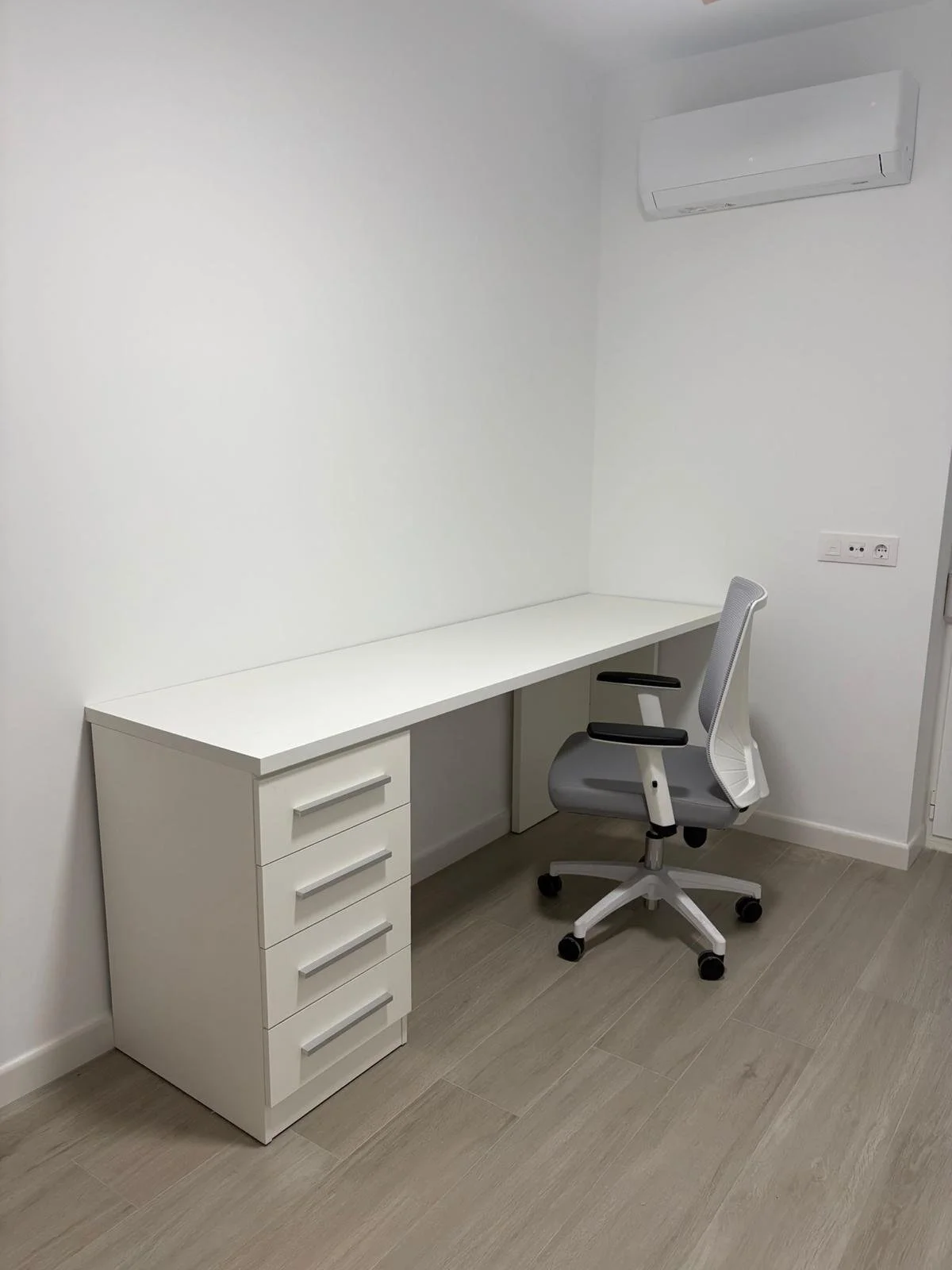 Escritorio blanco con gaveta, silla de oficina gris y blanca, aire acondicionado en la pared, enchufes eléctricos en la pared, suelo de madera, paredes blancas.