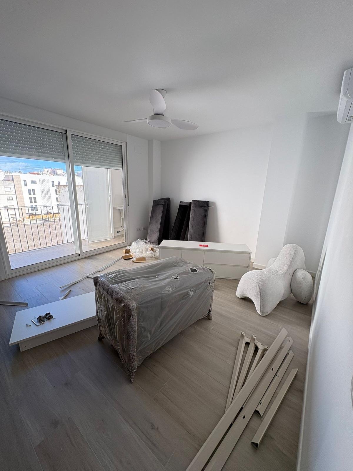 Sala moderna sin decorar con sofá cubierto y silla blanca con forma orgánica, vista de balcón con ciudad exterior, piso de madera clara, aire acondicionado y ventilador en techo.