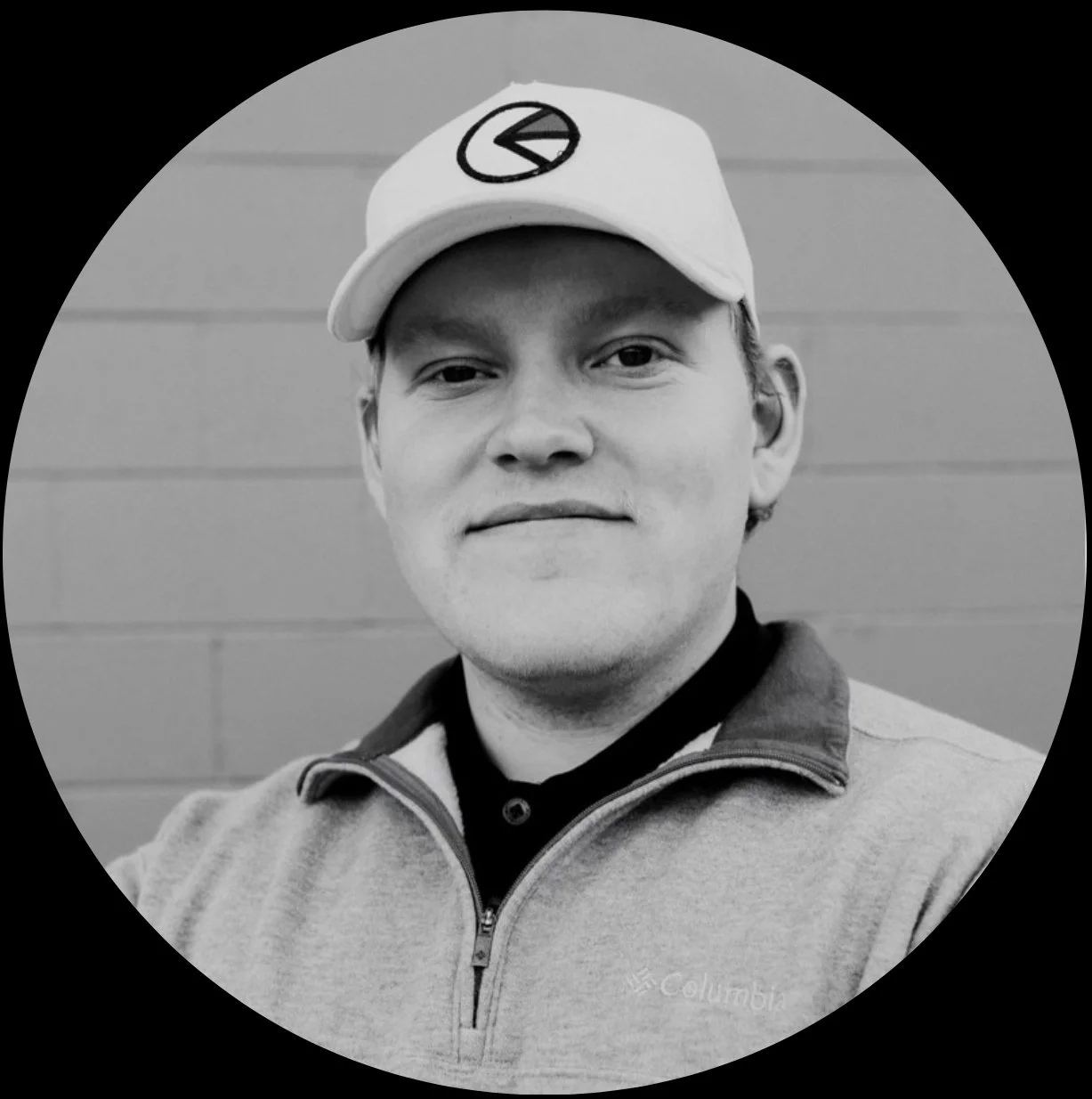 Black and white photo of a young man wearing a cap and zip-up jacket, standing in front of a wall.
