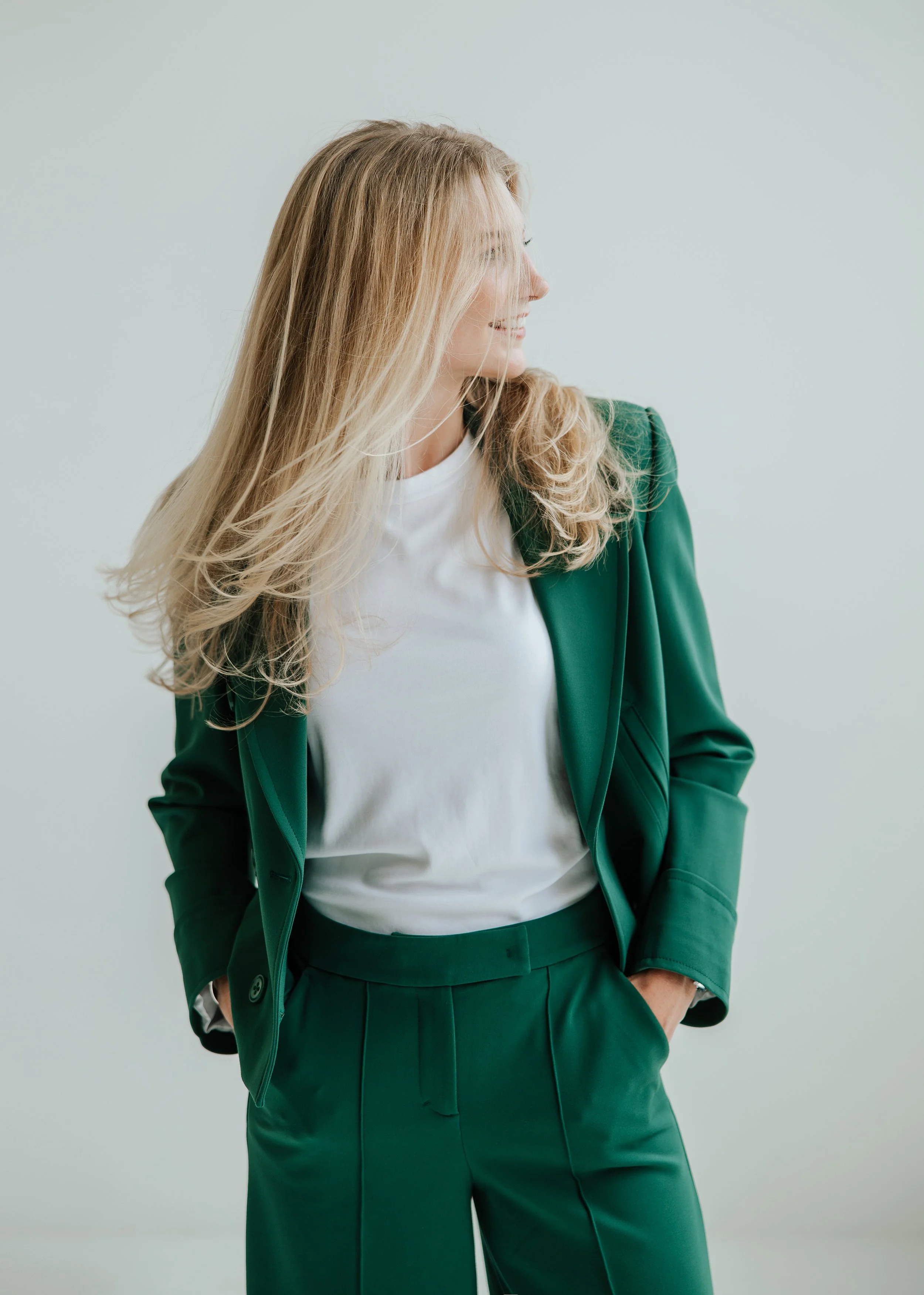 Eine Frau in einem grünen Anzug und weißem T-Shirt steht mit den Händen in den Taschen und schaut zur Seite.