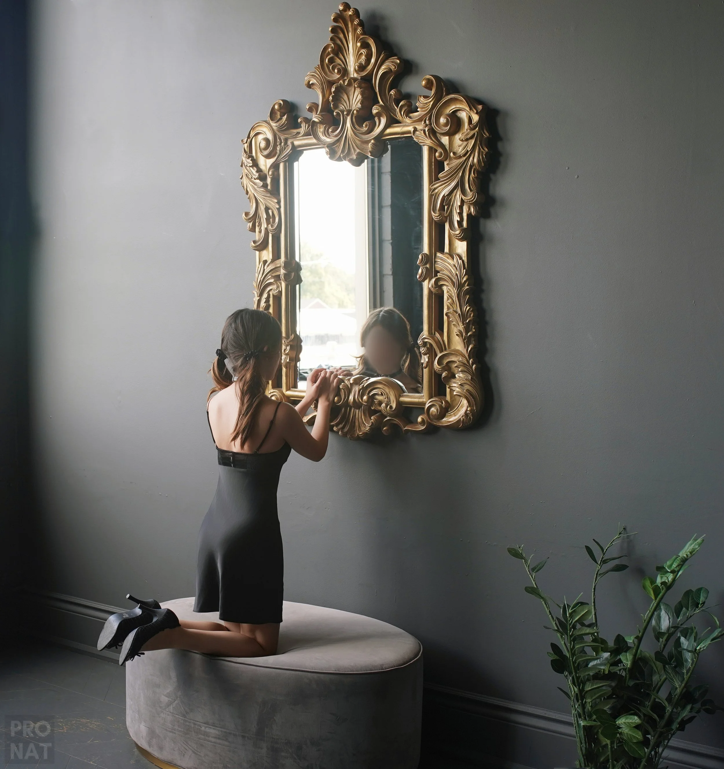 A woman kneeling on an oval ottoman, looking into a large ornate gold-framed mirror on a grey wall, with sunlight visible through a window reflected in the mirror and a green potted plant beside her.