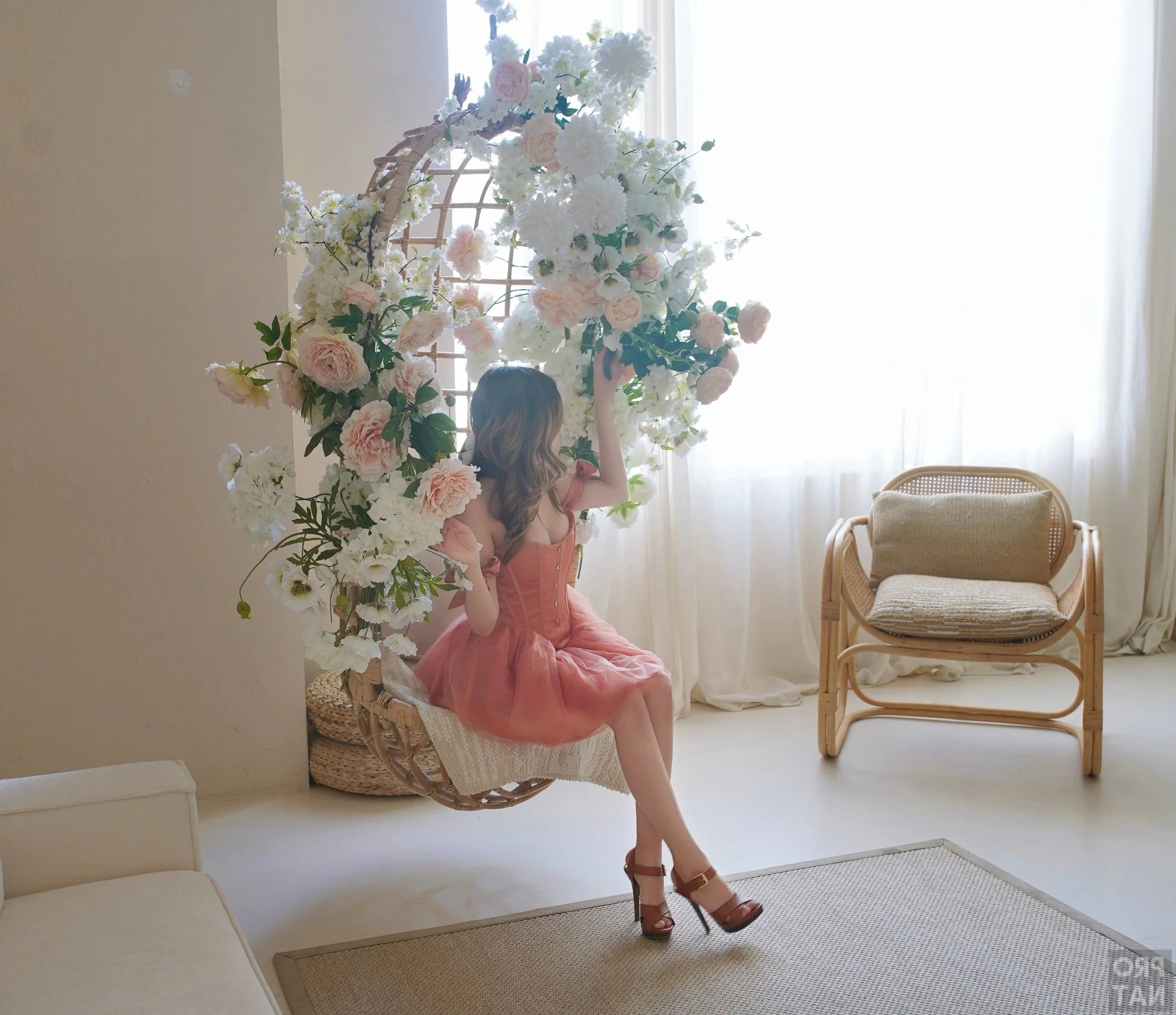 A woman in a peach-colored dress sitting on a hanging wicker swing decorated with white and pink flowers, in a bright, minimalist room with white curtains and furniture.