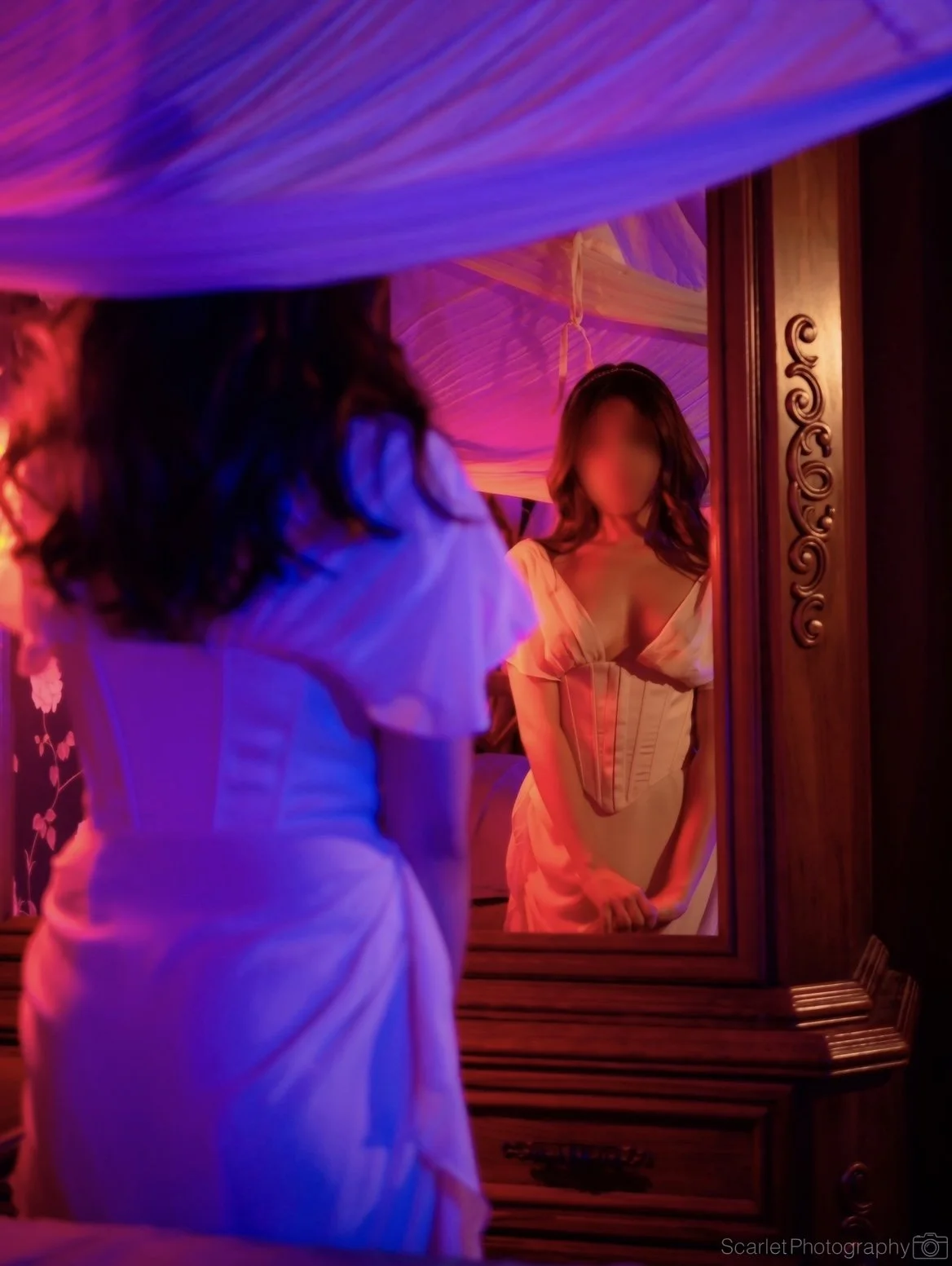 Woman in cream-colored dress looking at her reflection in a mirror surrounded by colorful, neon-like lighting.