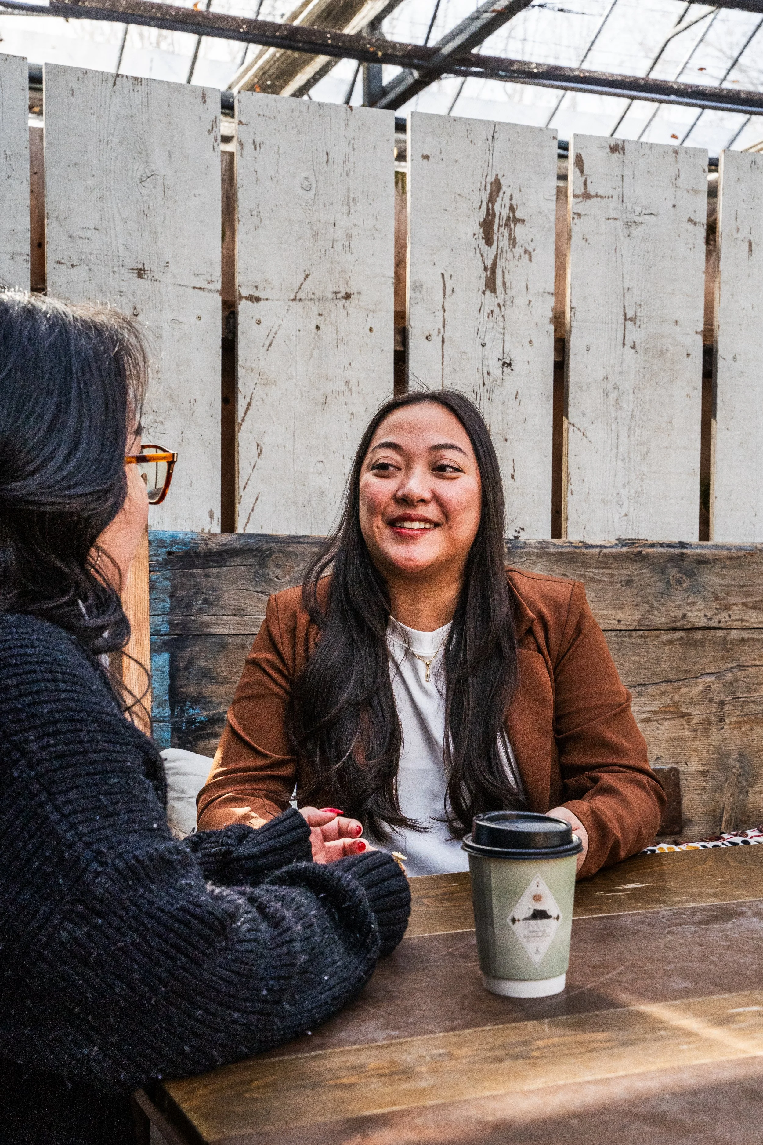Two women sitting at a wooden table having a conversation, one is wearing a black sweater and glasses, the other a brown blazer, a paper coffee cup is on the table.
