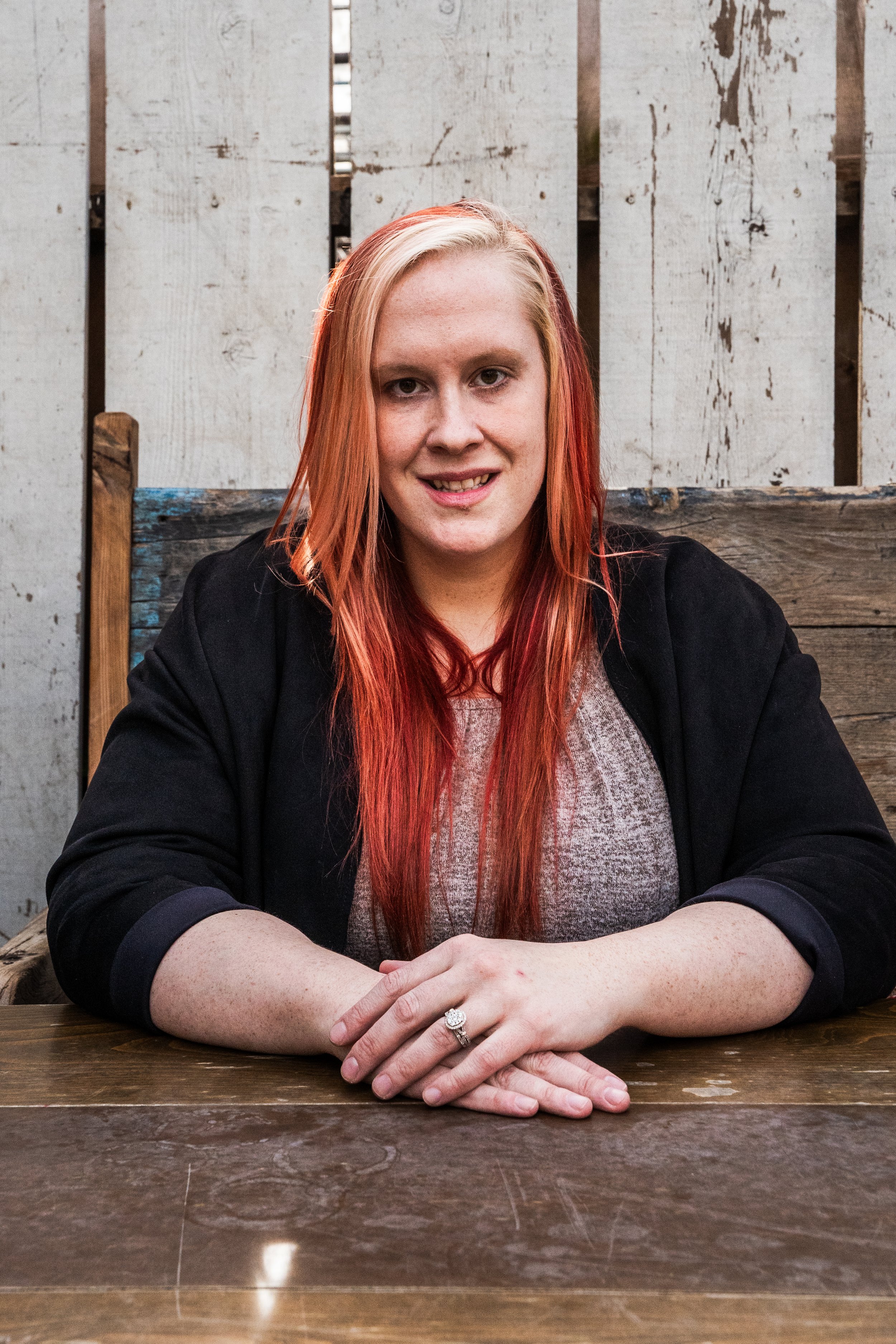 A woman with long, red and blonde hair sitting at a wooden table, smiling, with a wooden fence background.