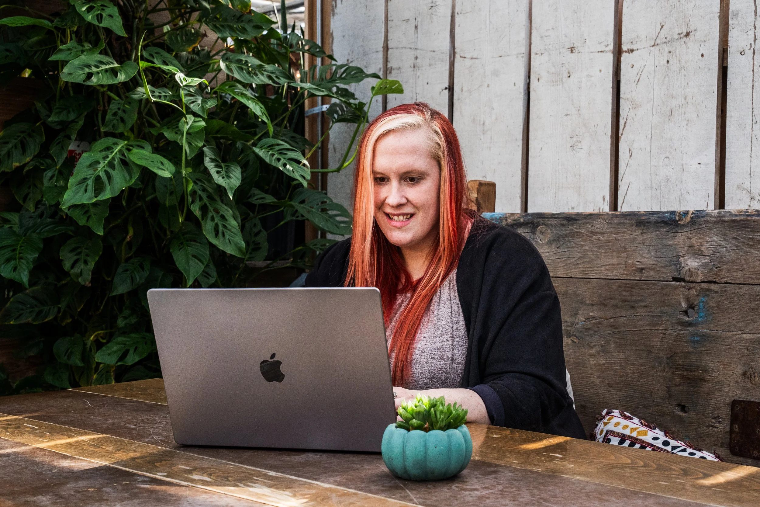 A woman with long red and blonde hair sitting at a wooden table, looking at a silver MacBook laptop, with green plants in the background and a small potted succulent plant on the table.