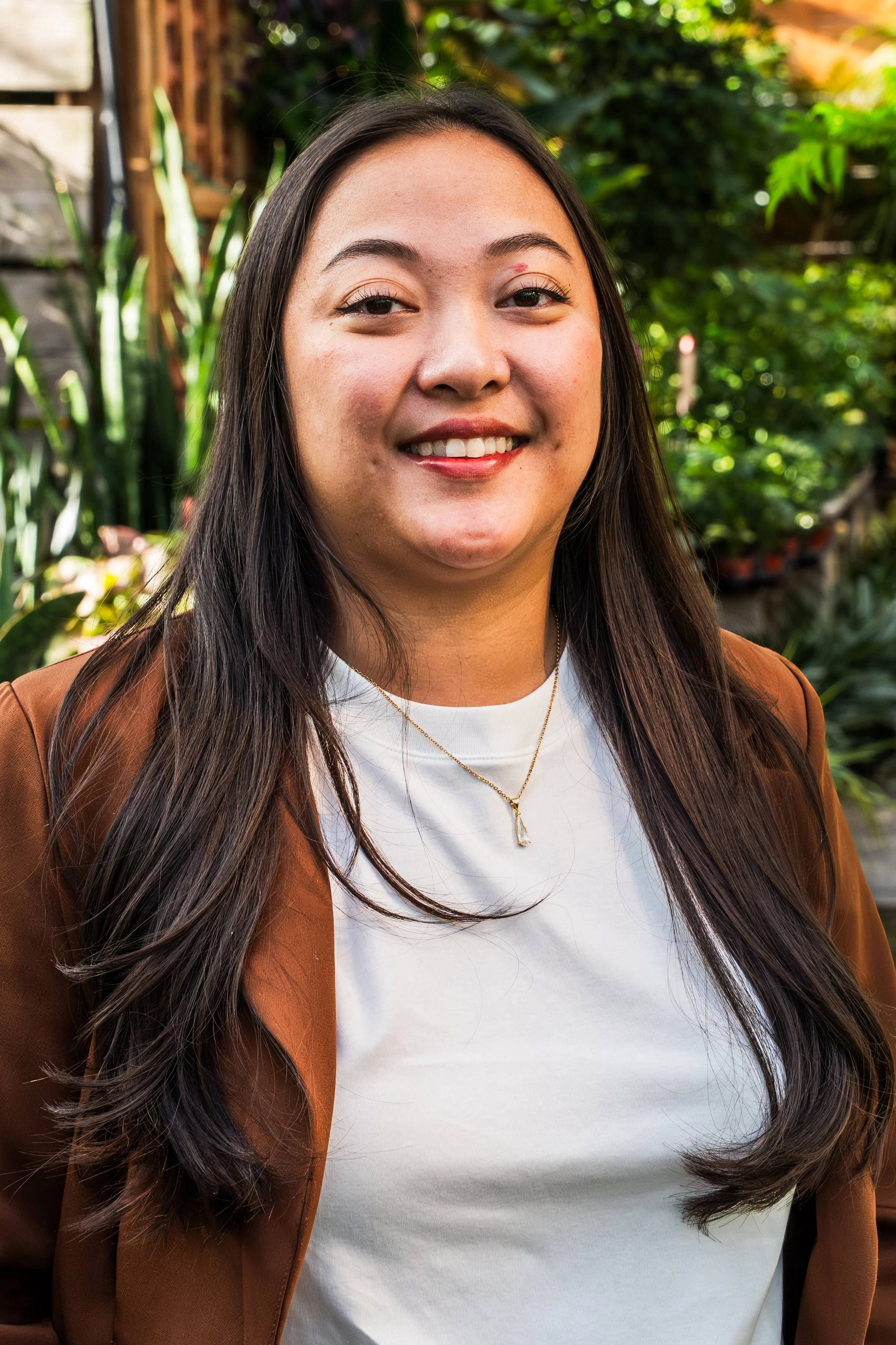 A young woman with long dark hair and a warm smile standing outdoors among green foliage, wearing a brown blazer, a white top, and a gold necklace.