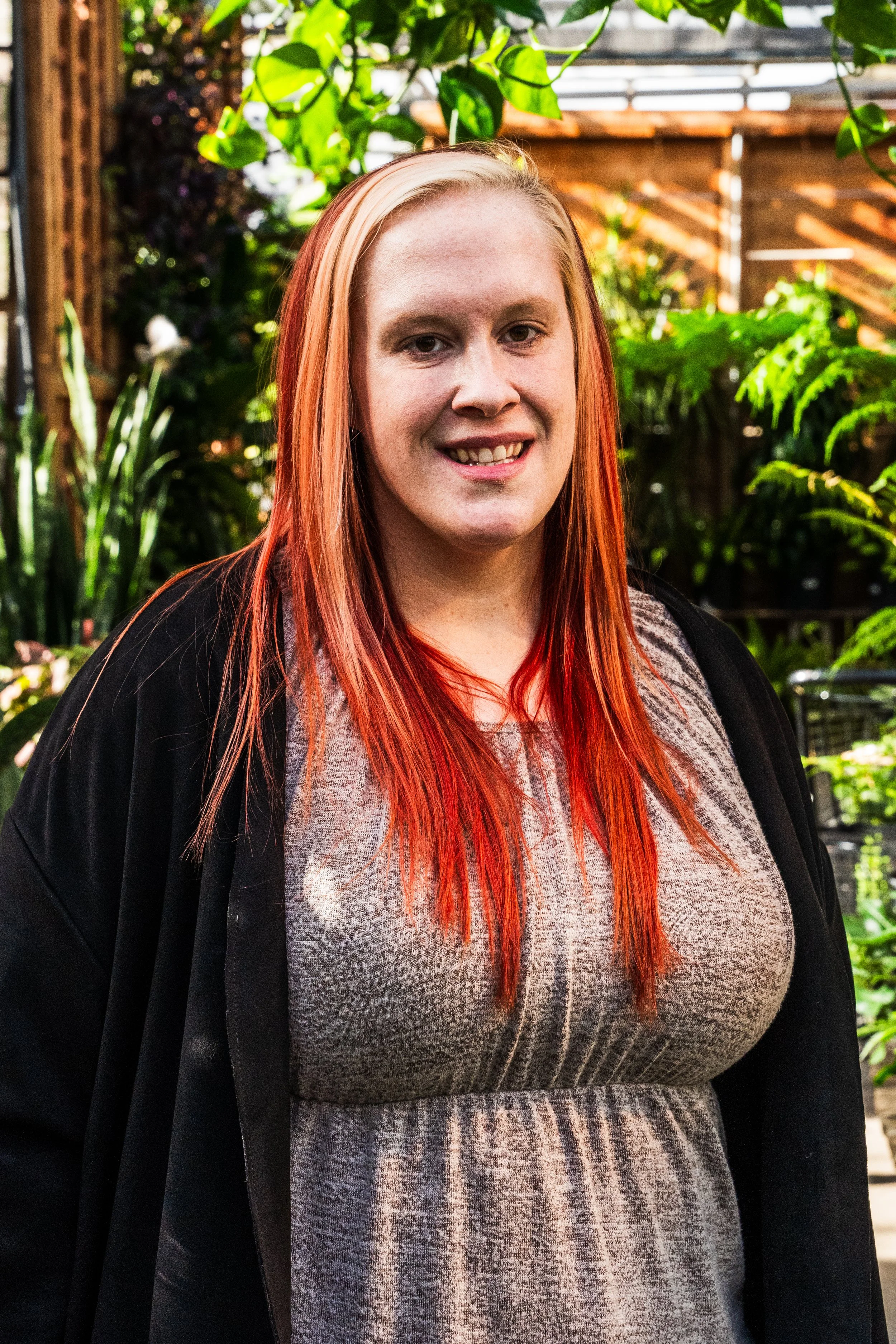 Young woman with long reddish hair smiling in a greenhouse or garden with lush green plants and wooden structures in the background.