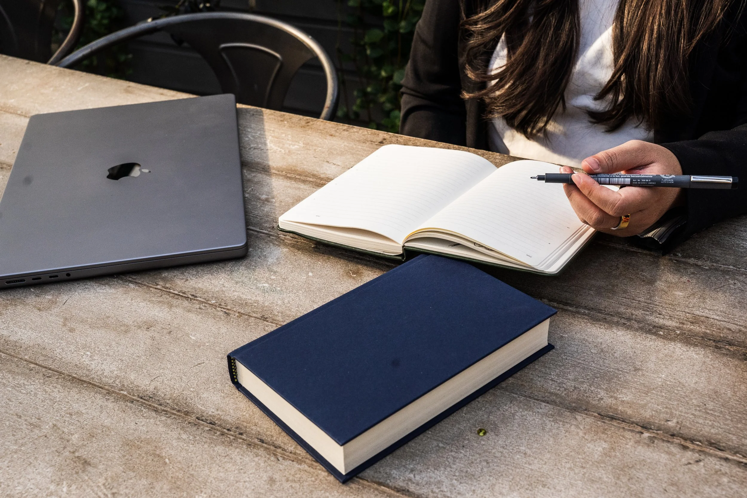 A woman writing in an open notebook with a pen, with a closed blue book, a closed laptop, and a black chair in the background on a weathered outdoor wooden table.