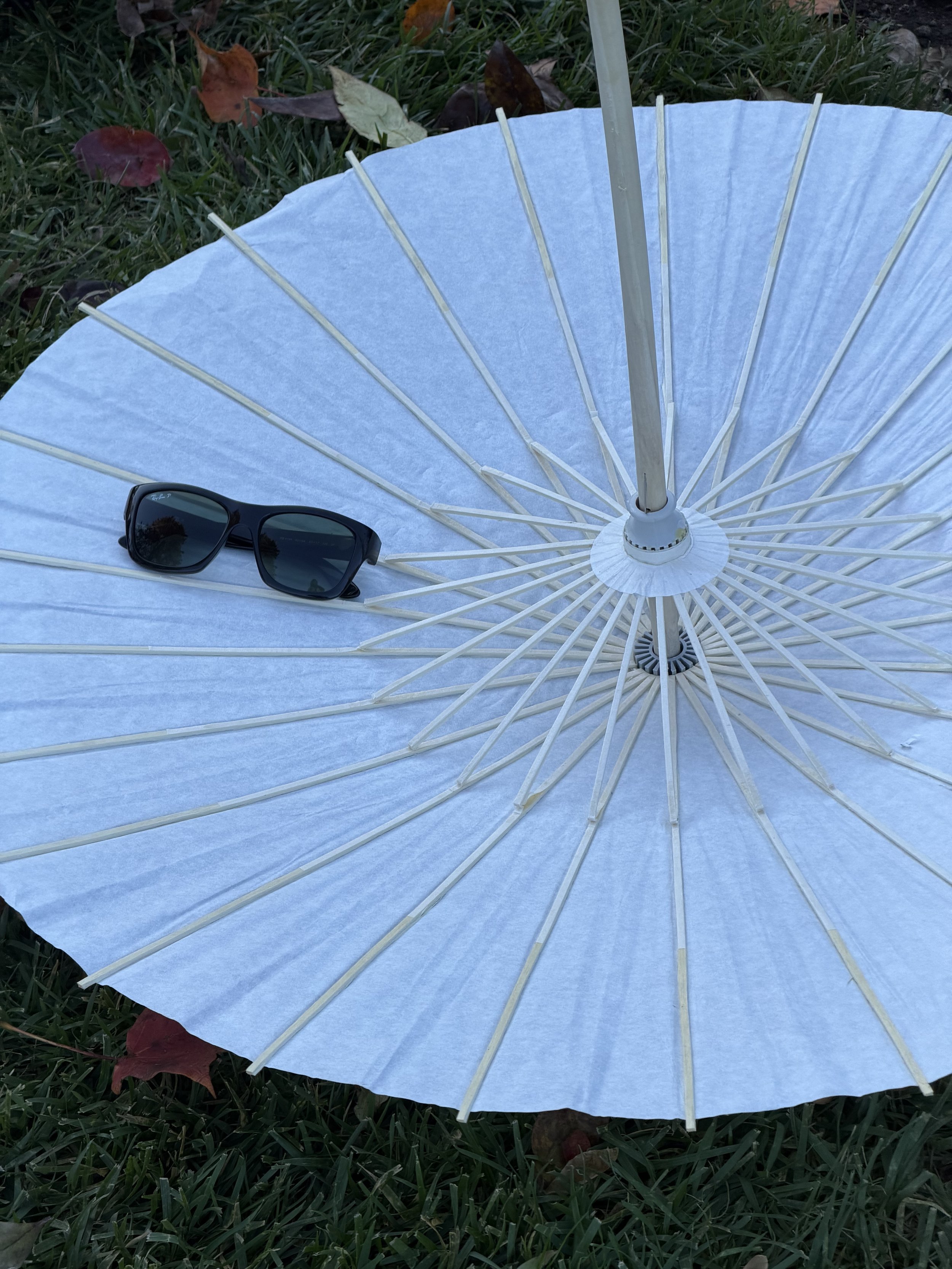 A white paper parasol with wooden ribs lying open on grass, with a pair of dark sunglasses resting on the parasol's surface.