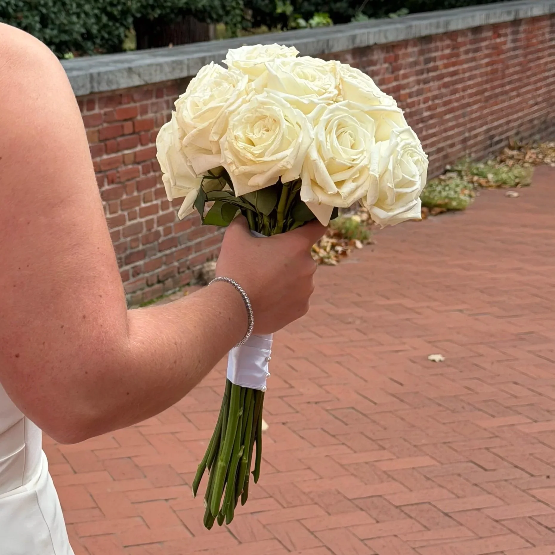 A person holding a bouquet of white roses with a brick wall and sidewalk in the background.