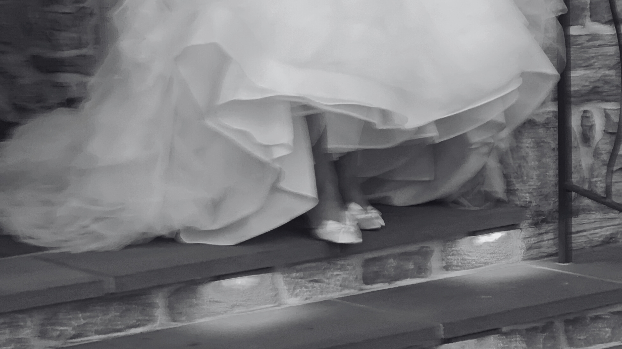 A person dressed in a wedding gown standing on a wooden stage or platform, with a lace or embroidered train and high-heeled shoes. The background appears to be a rustic brick wall.