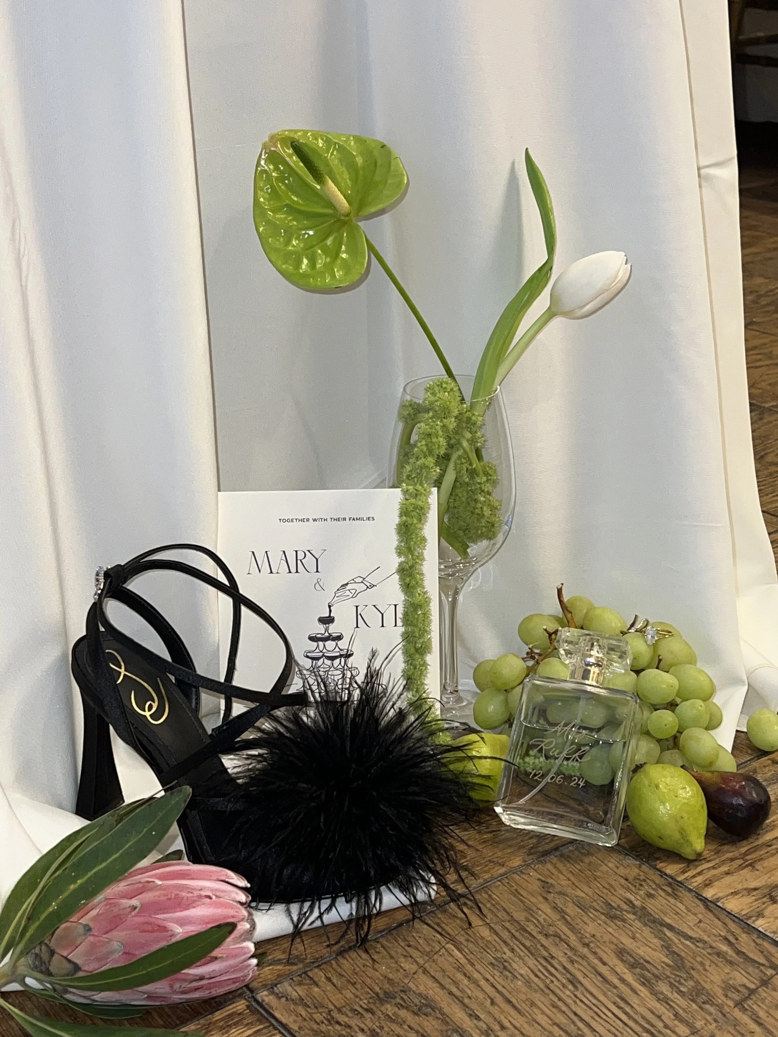 A floral arrangement with green leaves, white tulips, and green moss in a glass vase, accompanied by a black feathered accessory, green grapes, a perfume bottle, and a pink flower on a wooden table.