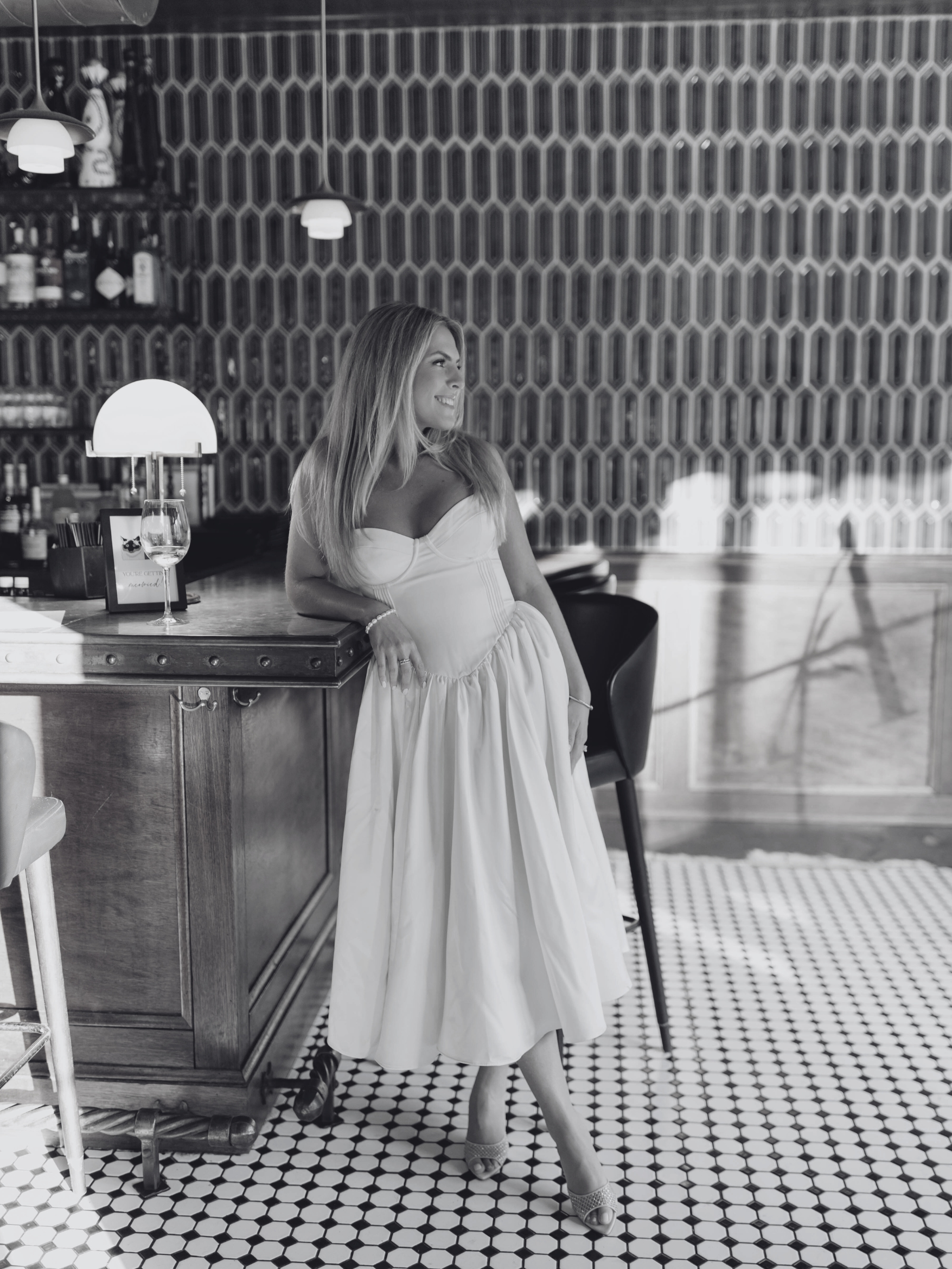 A woman in a dress standing at a bar with a glass of wine, in a stylish interior with patterned wall and tiled floor.