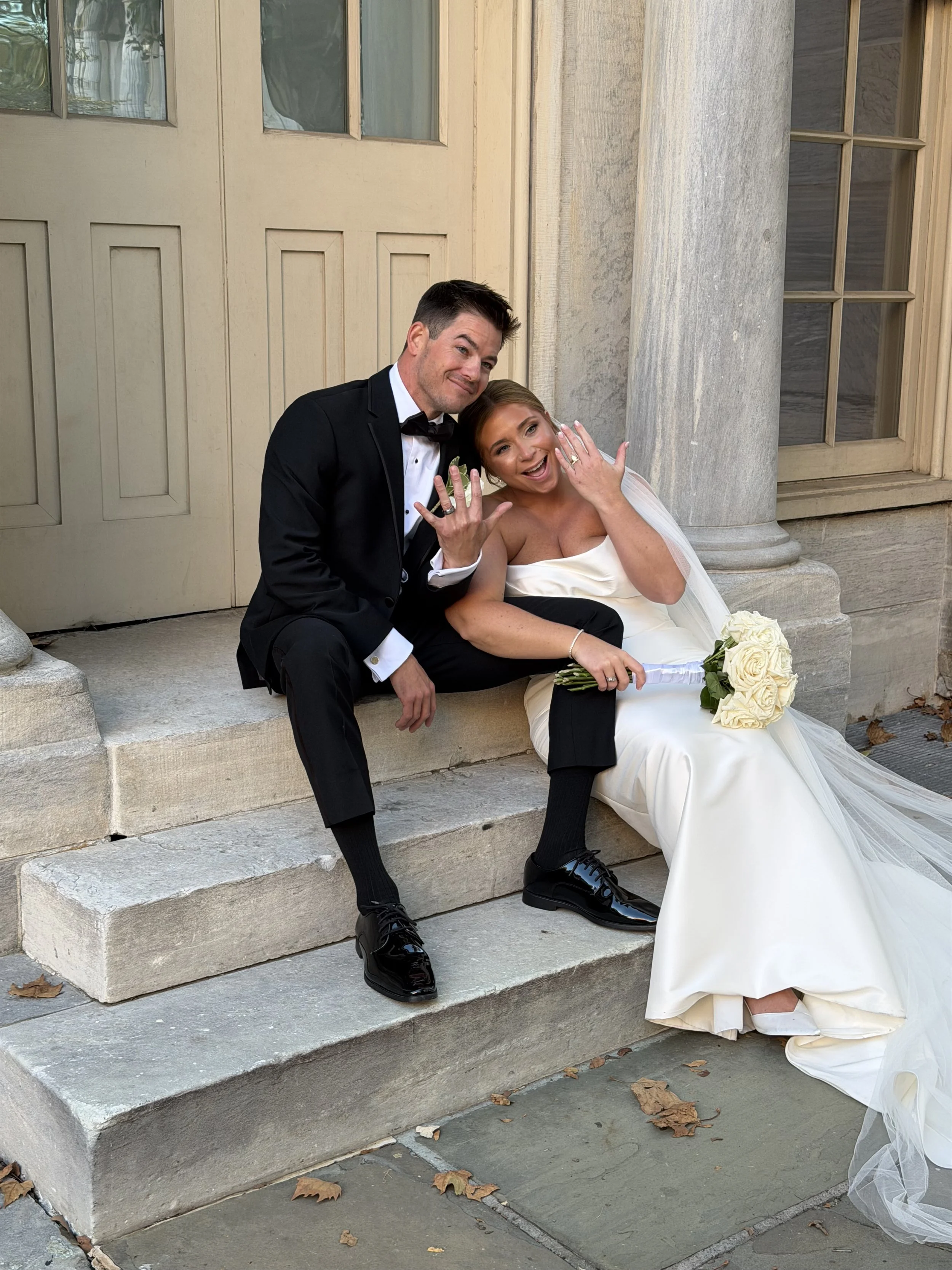 A newlywed couple sitting on the steps outside a building, showing off their wedding rings and smiling happily, with the bride holding a bouquet of white roses.