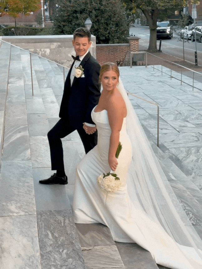 A newlywed couple in wedding attire, standing on marble steps outside, holding hands and smiling. The bride is holding a bouquet of white flowers and wearing a white strapless wedding gown with a long train and veil. The groom is dressed in a black tuxedo with a bow tie and a white boutonniere.