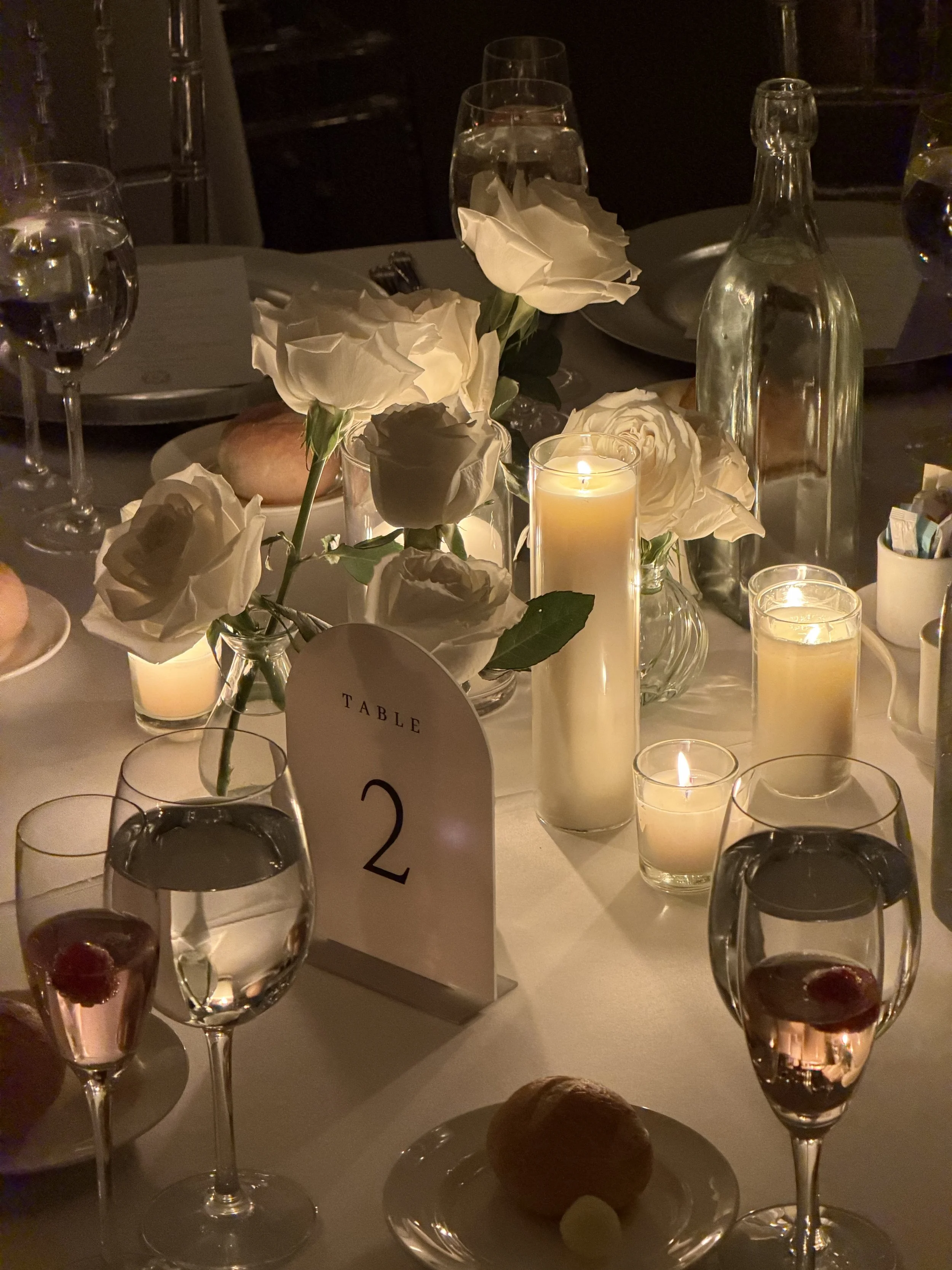 A elegantly set dining table with white roses and candles, glasses of water and wine, a table number 2 sign, and plates with bread rolls. The lighting is dim and warm.