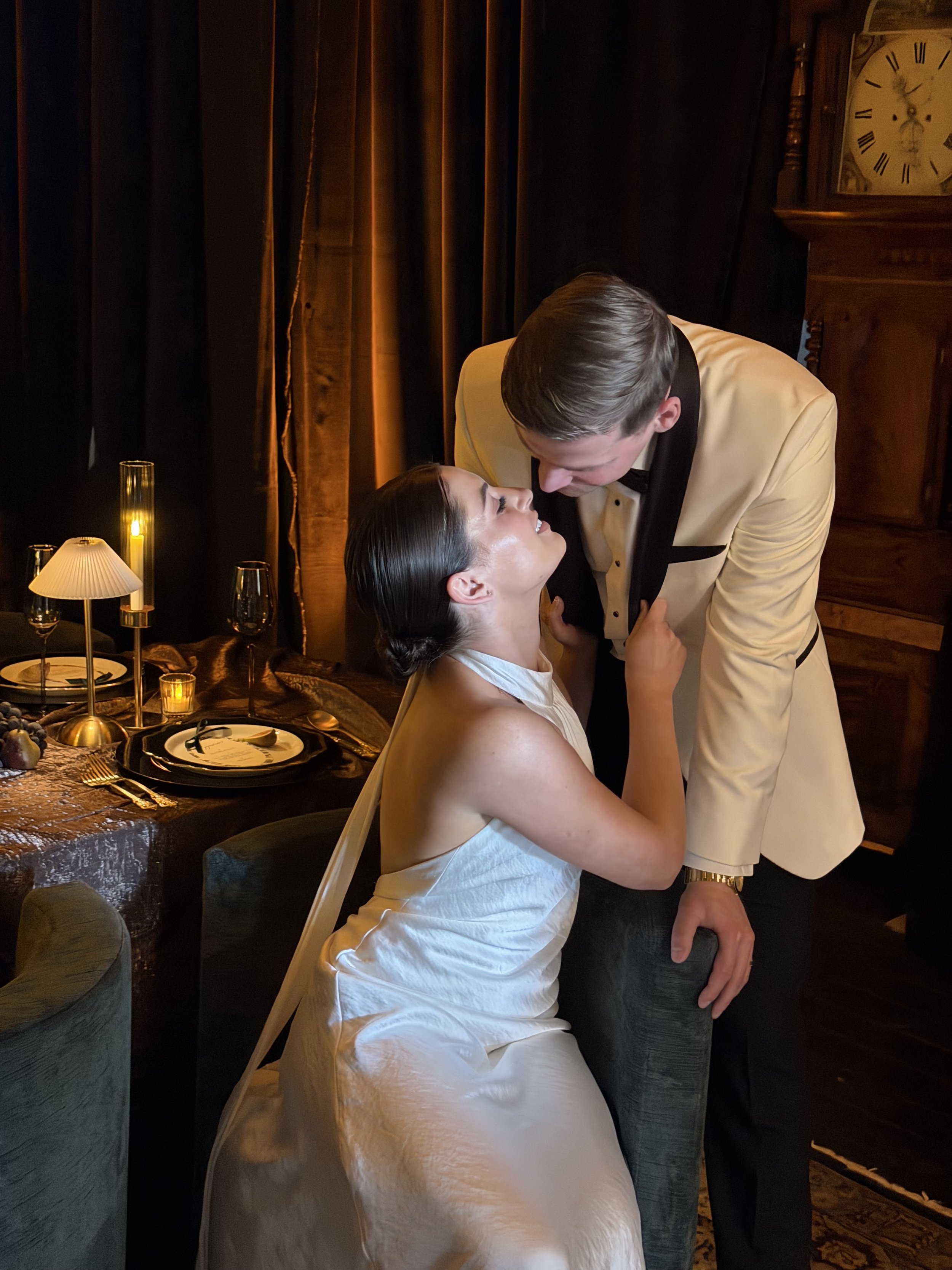A couple dressed in formal attire in an intimate moment at a dinner table, with the woman sitting and the man leaning over her in a romantic pose.