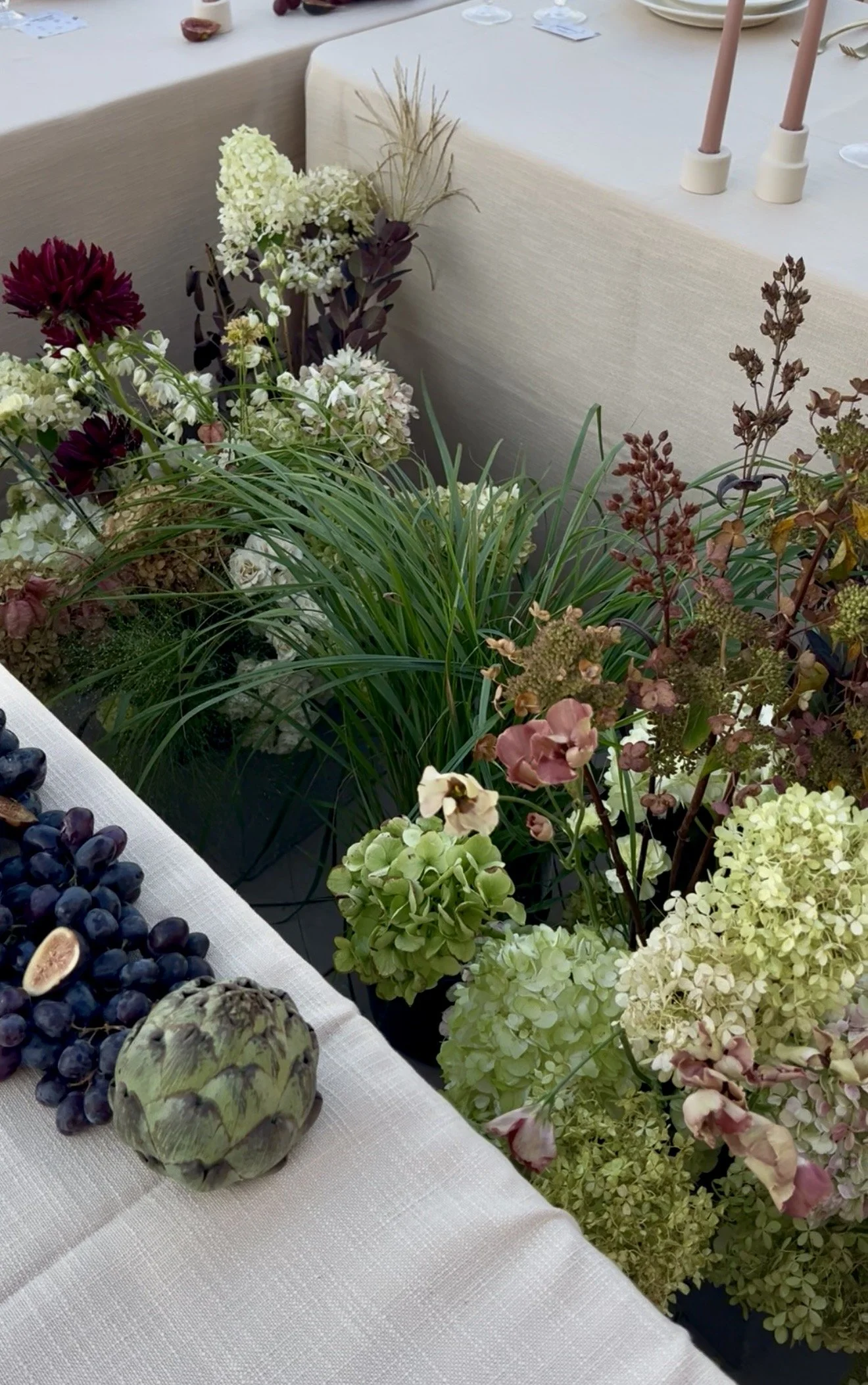 A table decorated with an assortment of green and white hydrangeas, purple and pink flowers, green foliage, a cluster of dark grapes, and a small artichoke. The background includes a beige tablecloth with candles and plates.