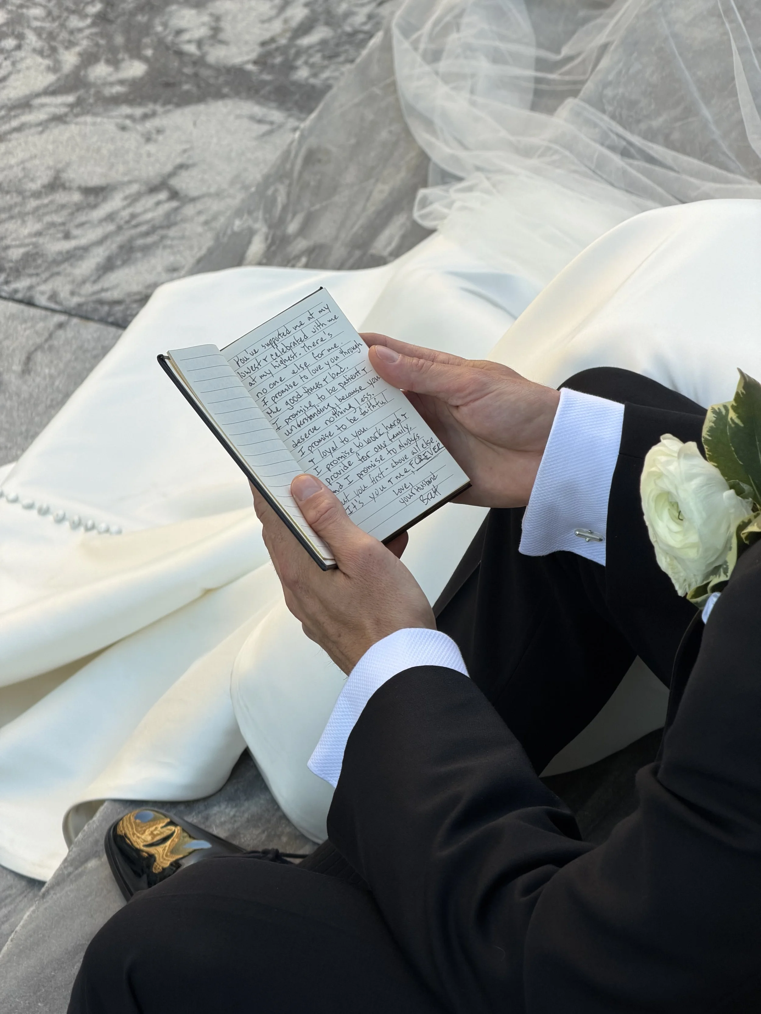 Person in formal wedding attire sitting on a stone surface, holding and reading a handwritten note in a small notebook. The person is wearing a black suit with white shirt cuffs, and a white boutonniere is attached to their lapel. A white wedding dress and veil are visible on the ground beside them.