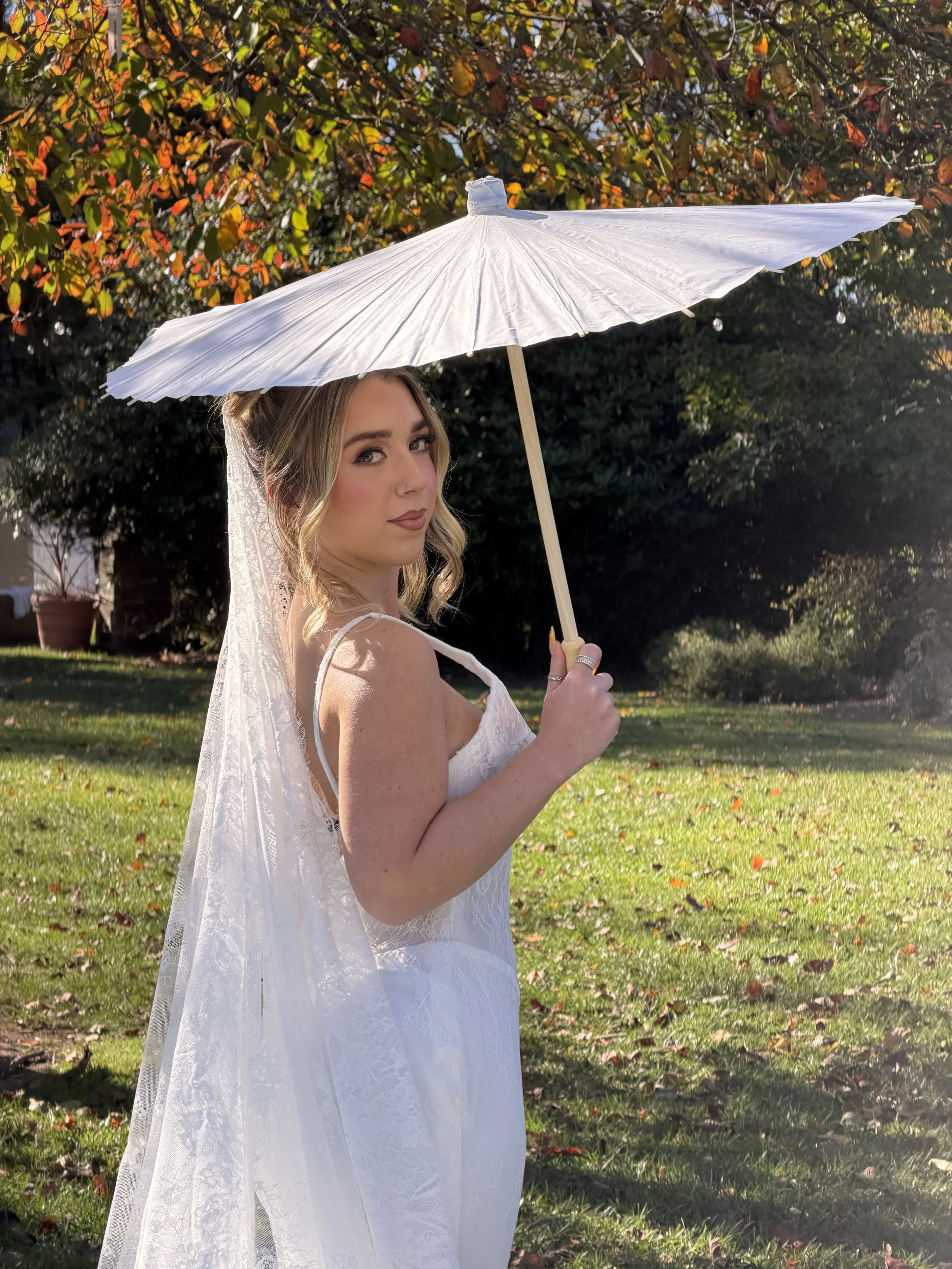 Woman in white dress holding a white parasol outdoors in a park on a sunny day.