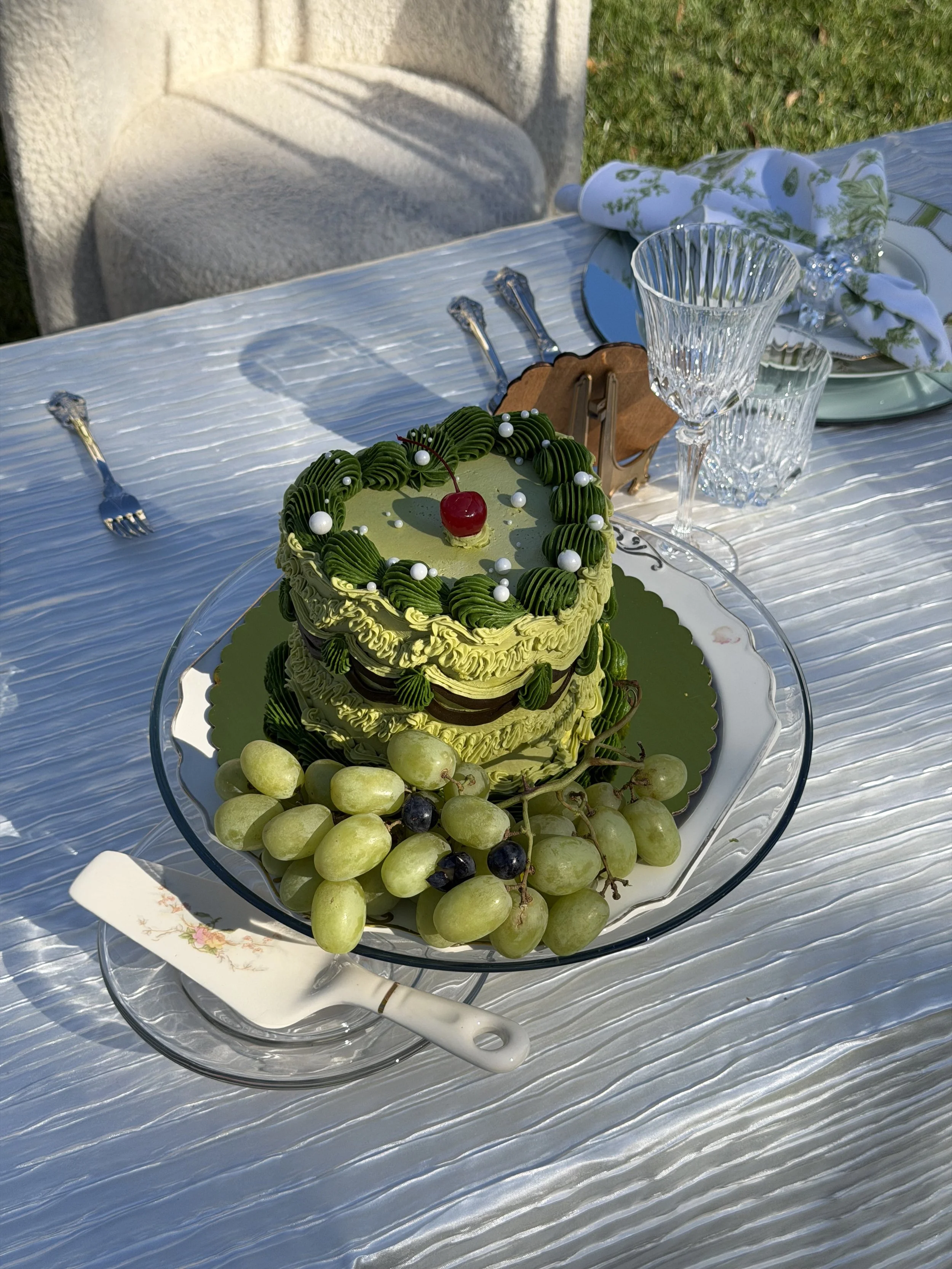 A green layered cake with decorative cream, topped with a cherry and white pearls, surrounded by green grapes on a glass platter, set on an outdoor table with crystal glasses, plates, and silverware.