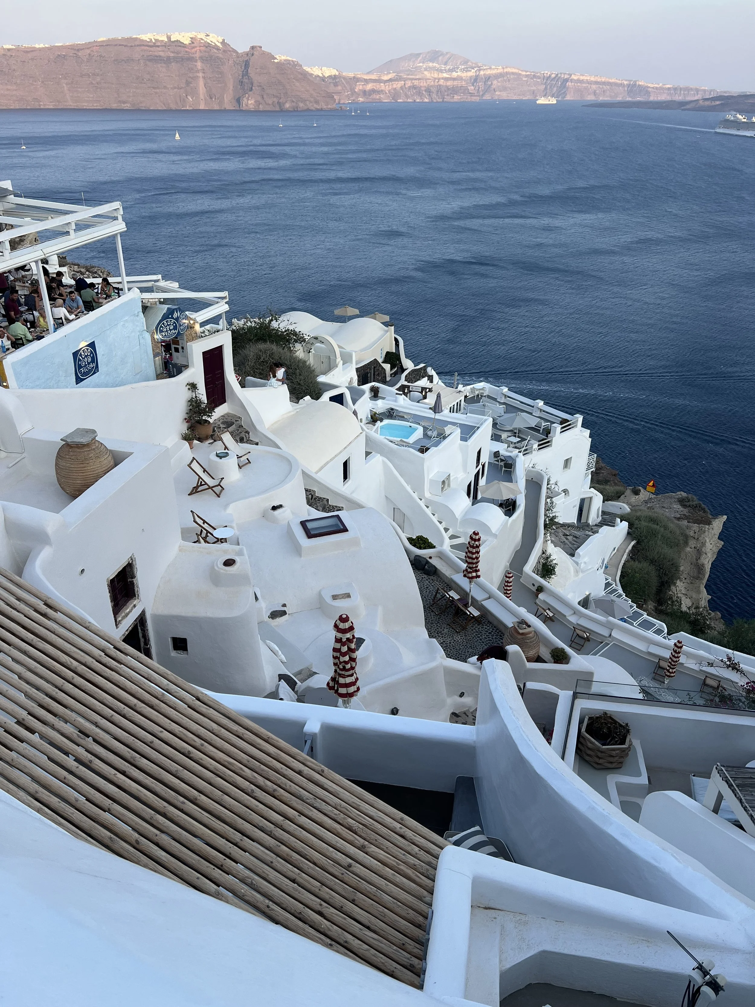 A scenic view of white-washed buildings with terraces and pools on a hillside overlooking the deep blue Aegean Sea, with sailboats and a volcanic island in the background.