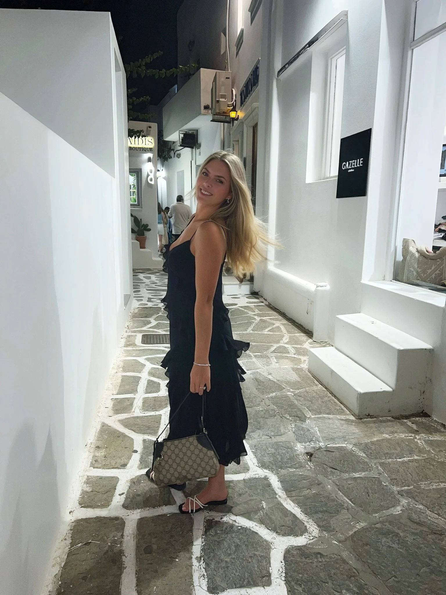 A young woman with long blonde hair smiling while standing on a stone pathway outside a white building at night. She is wearing a black dress, black high heels, and carrying a beige patterned handbag.