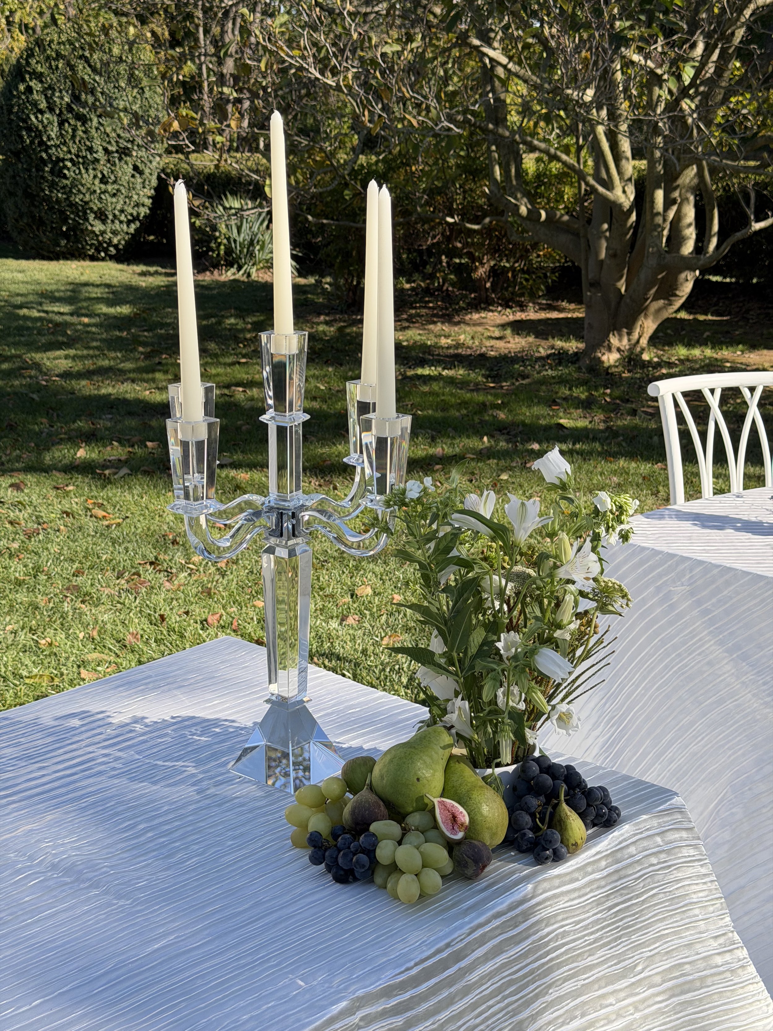 Table decorated with a glass candelabrum, a bouquet of white flowers, and an assortment of green and purple grapes, pears, and a halved fig, set outdoors on a white tablecloth with green grass and trees in the background.
