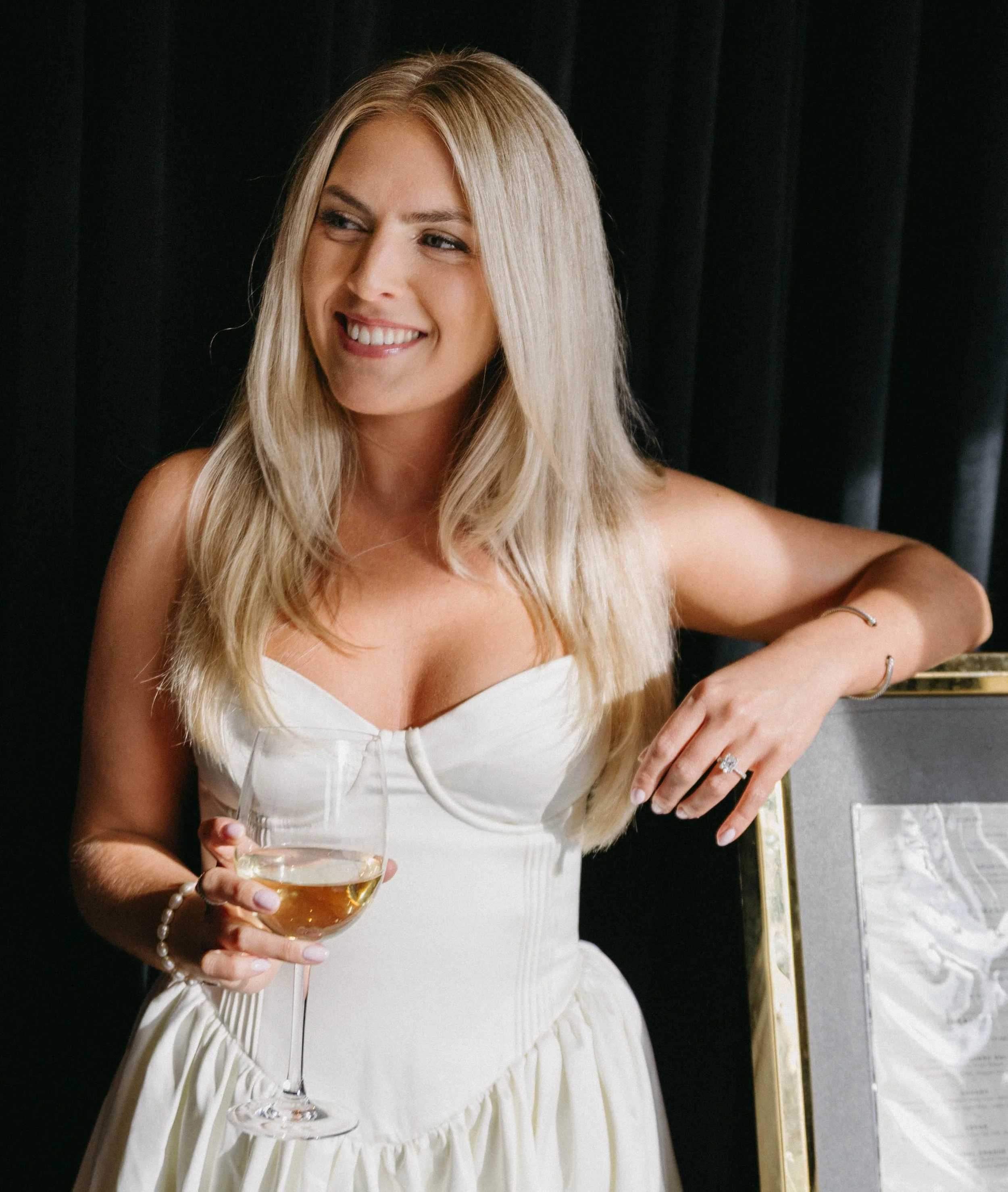 A woman with blonde hair smiling, wearing a white dress, holding a glass of white wine, standing next to a framed picture, dark background.