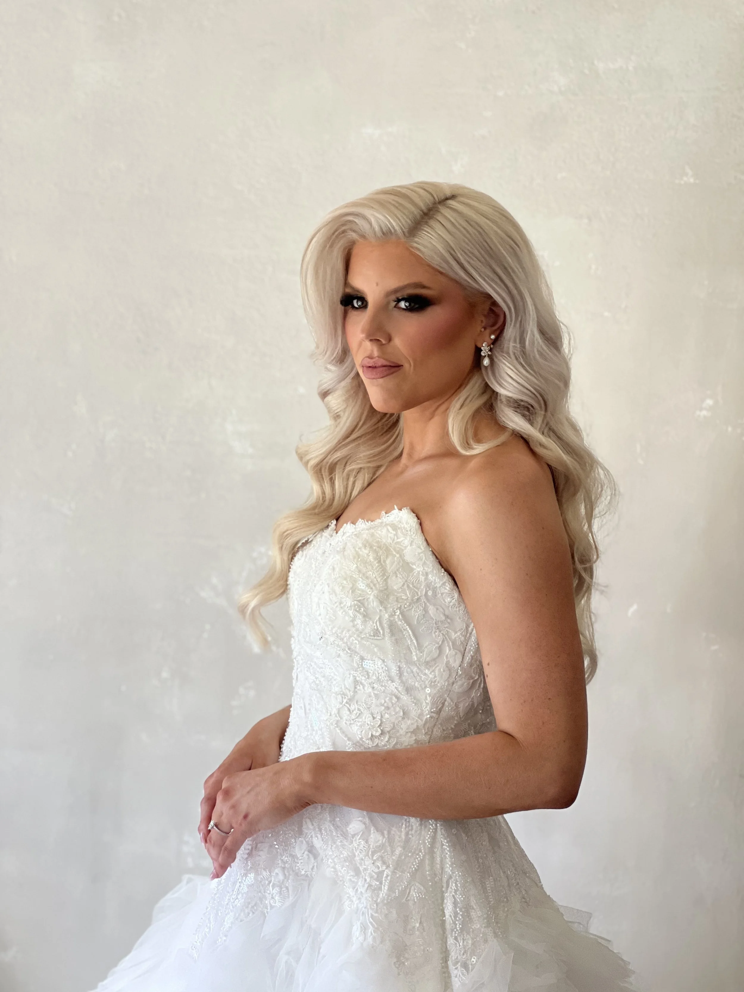 A woman in a white wedding dress with lace details, sporting long blonde hair in soft waves, with makeup and earrings, standing against a plain light-colored background.