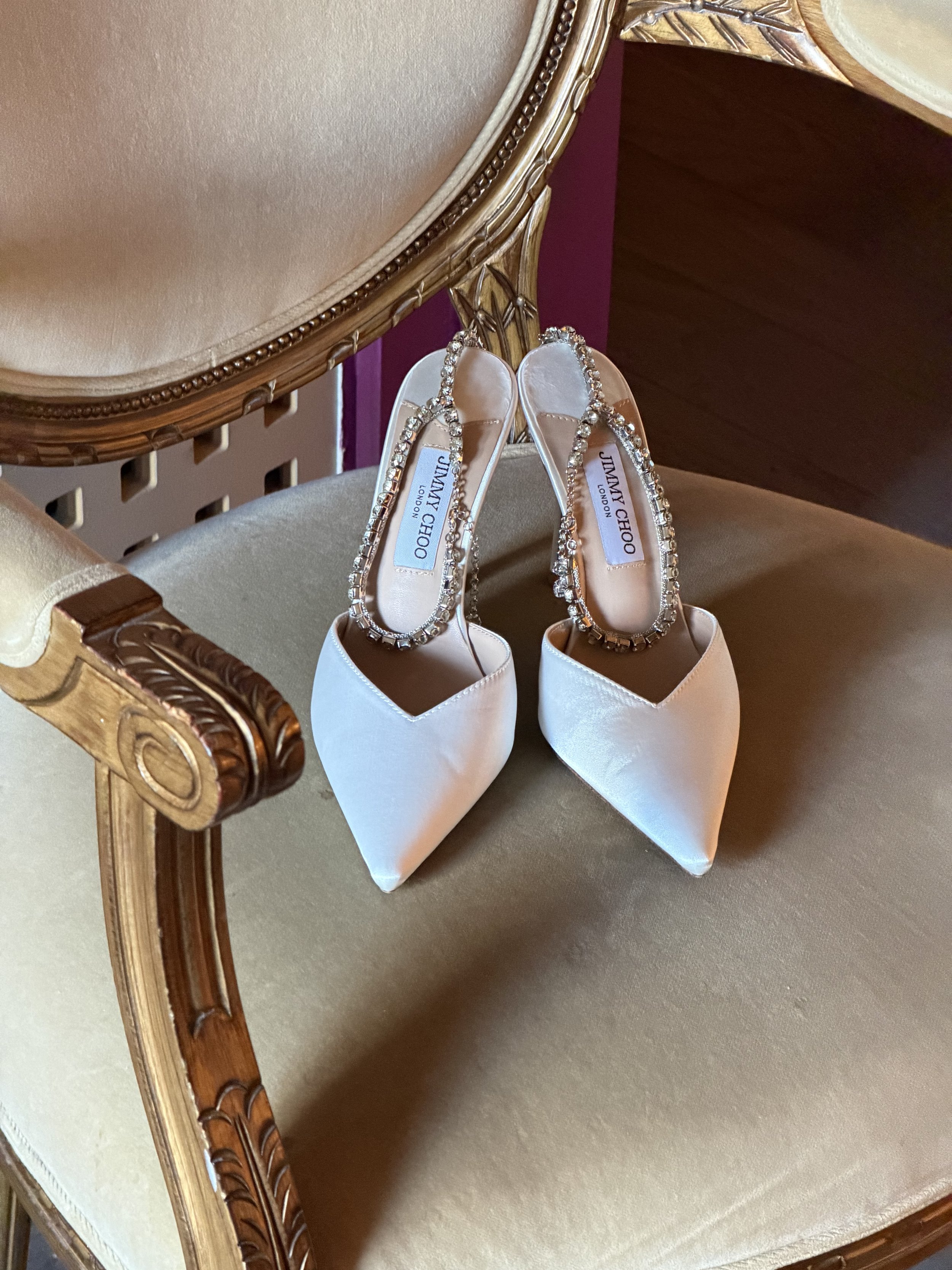 A pair of white satin Jimmy Choo high heels with decorated straps, placed on a vintage-style beige upholstered chair.