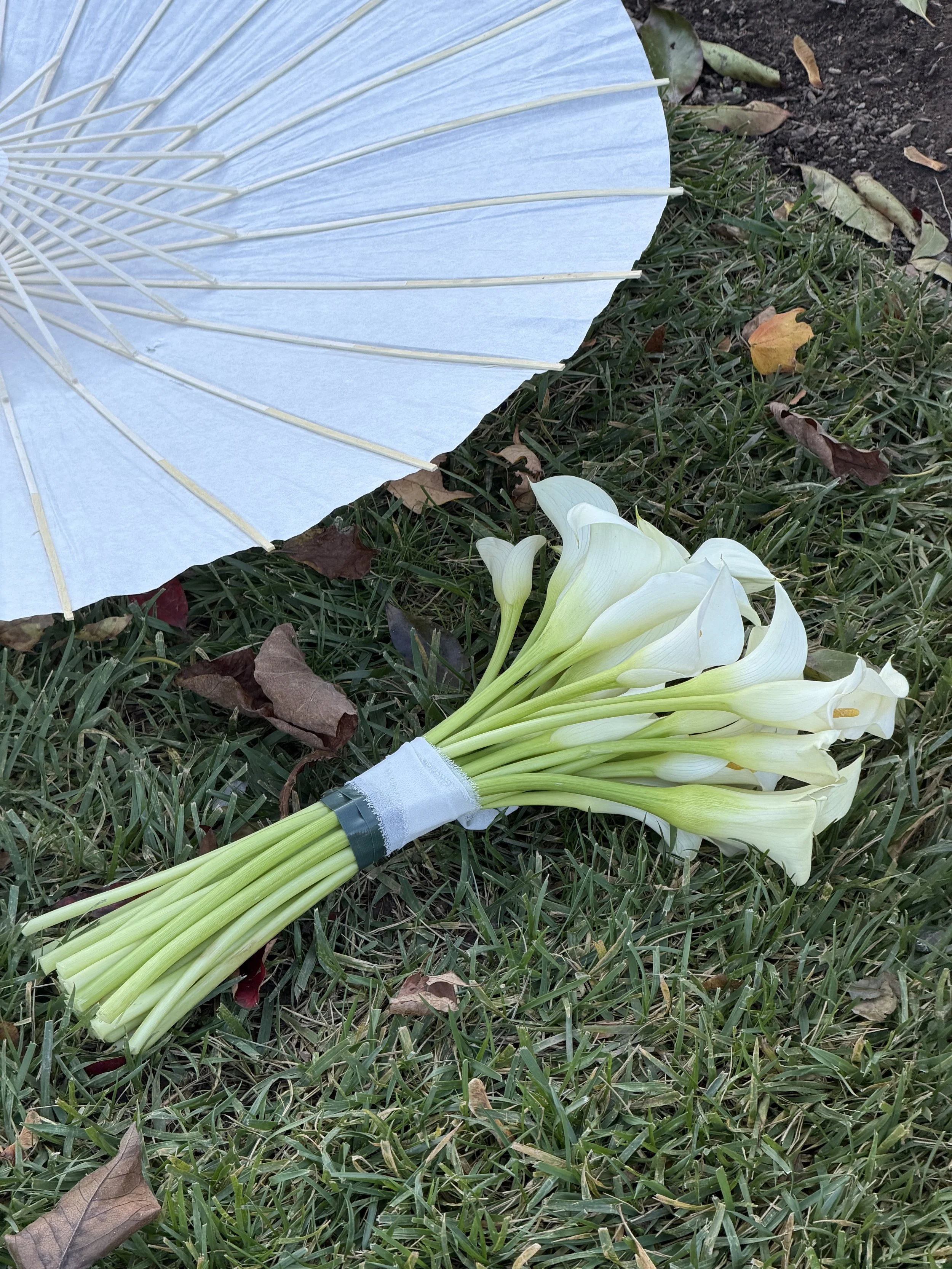 A white parasol and a bouquet of white calla lilies on the grass with fallen leaves.