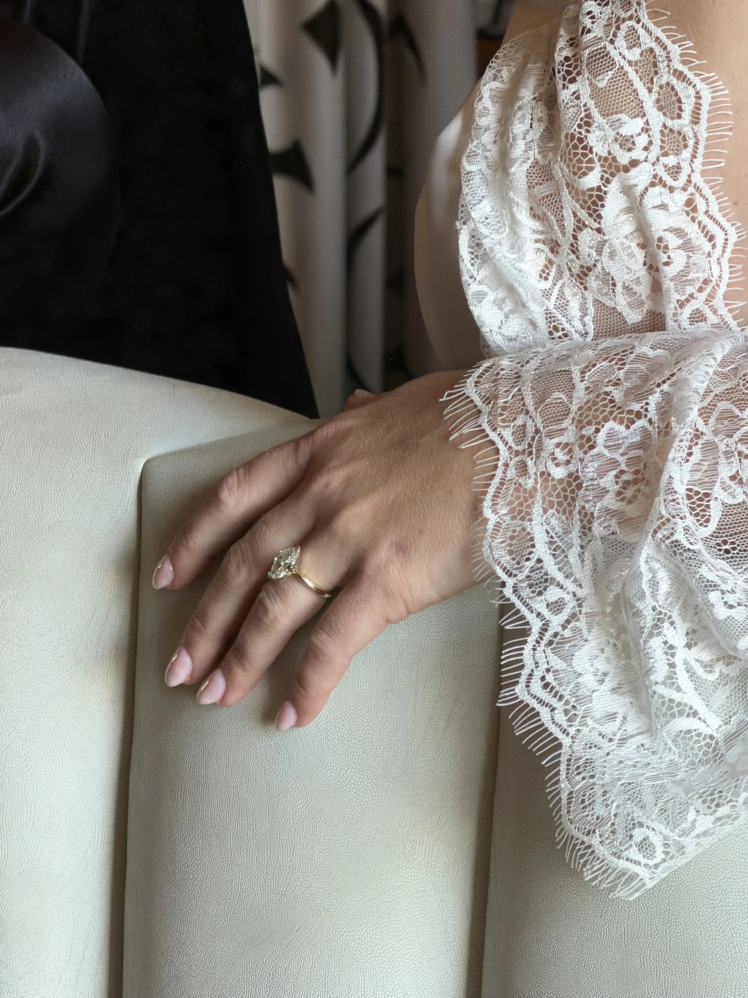 Close-up of a woman's hand resting on a beige leather surface, wearing a gold ring with a large gemstone, and dressed in a white lace gown with scalloped edges.