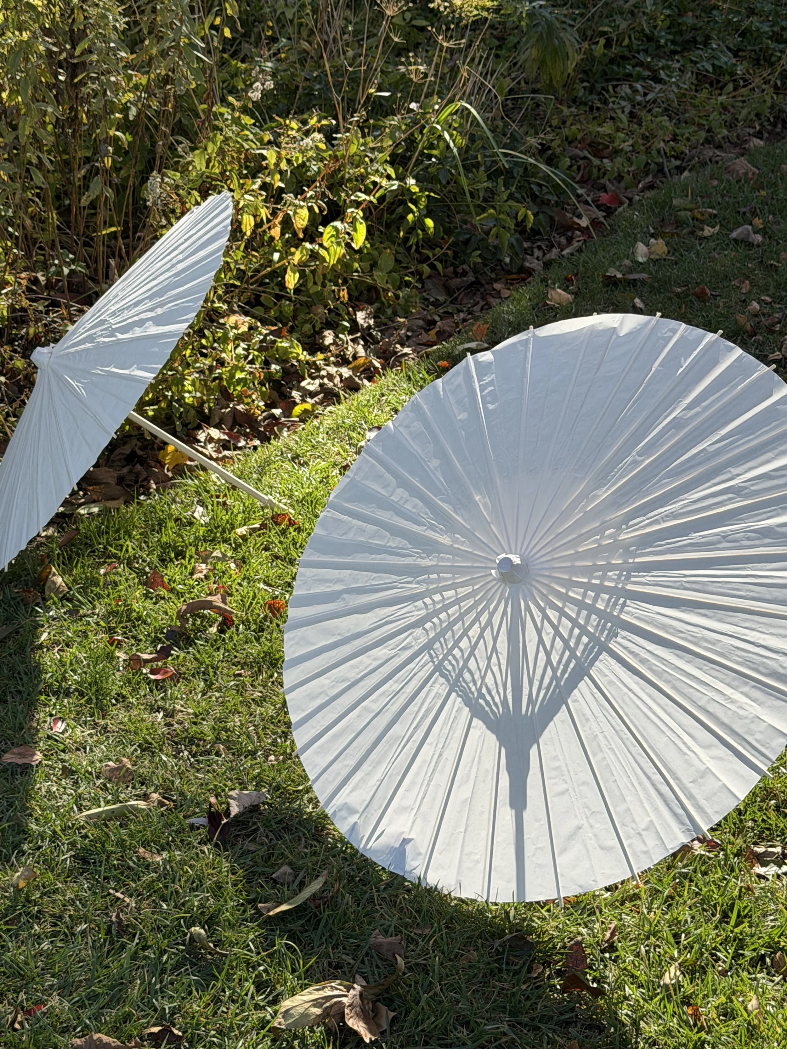 Two white paper parasols, one open and casting shadows, outdoors on green grass with fallen leaves and bushes in the background.