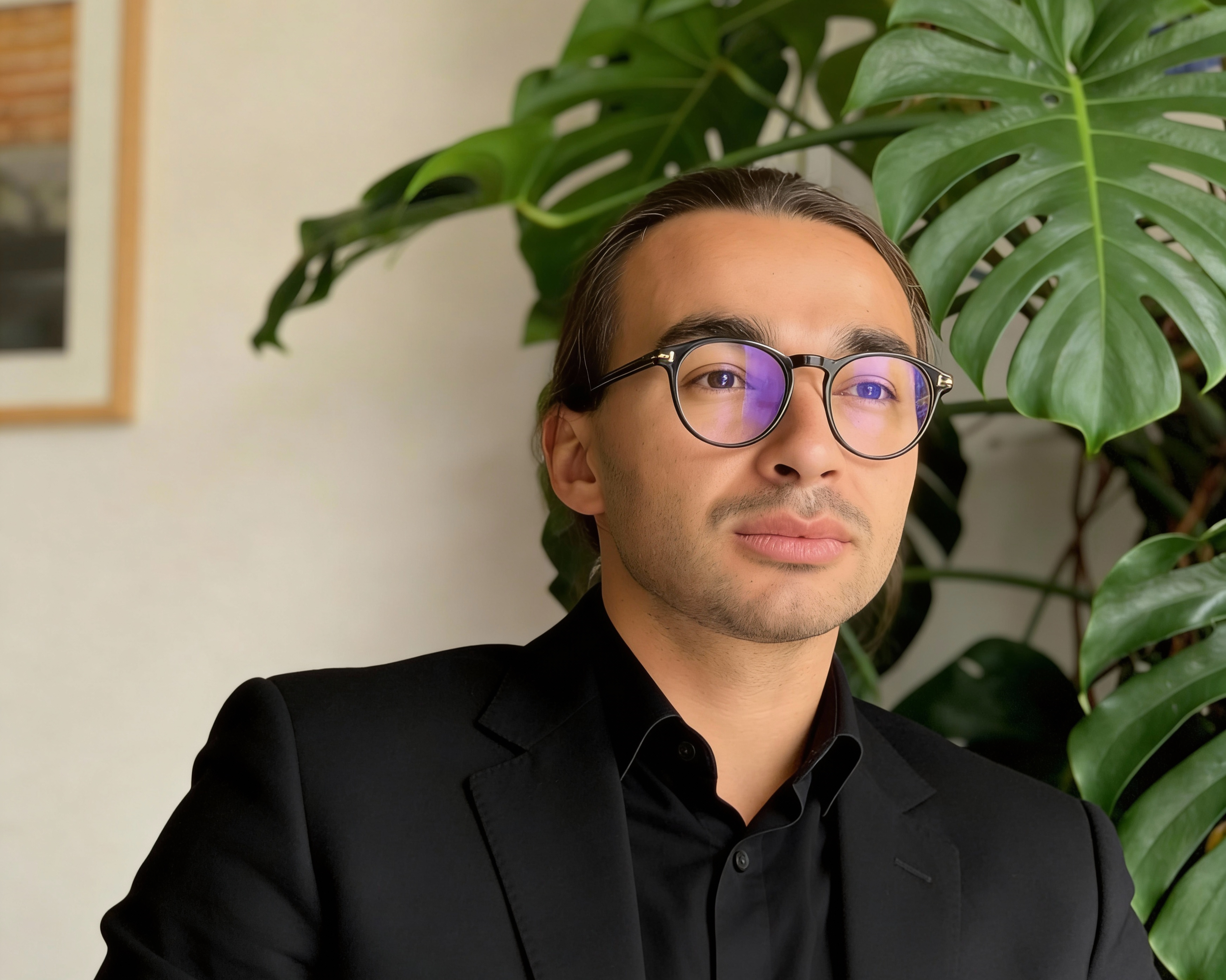 Un homme portant des lunettes et un costume noir, assis devant un grand plante verte dans un intérieur.