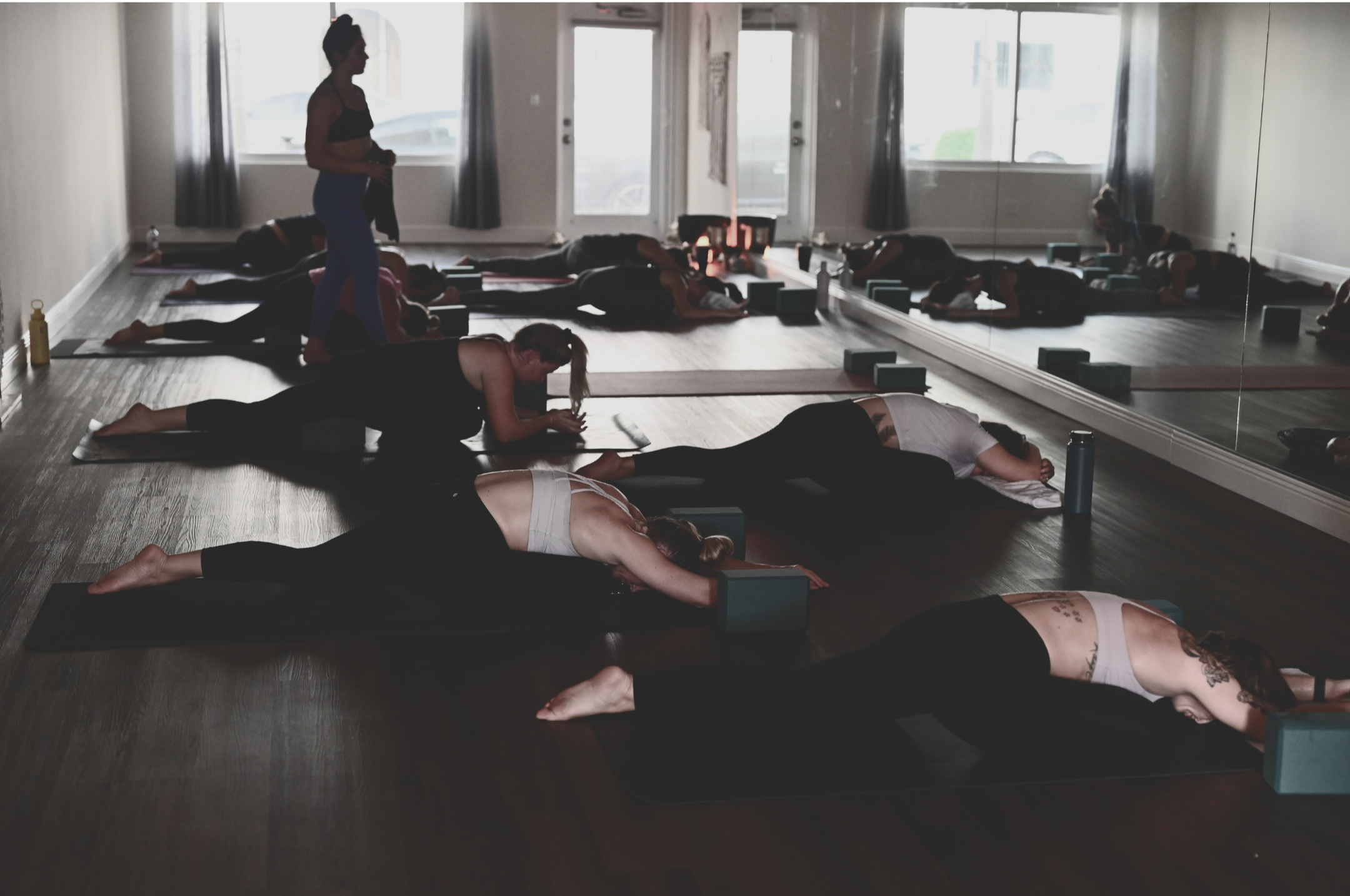 Brittney Coates Yoga YogaUX, user-friendly yoga for your inner space. yoga workshop for arm balances and invers-group of people doing a yoga class in a studio with mirrored wall, lying on yoga mats in a restorative pose, with an instructor standing.