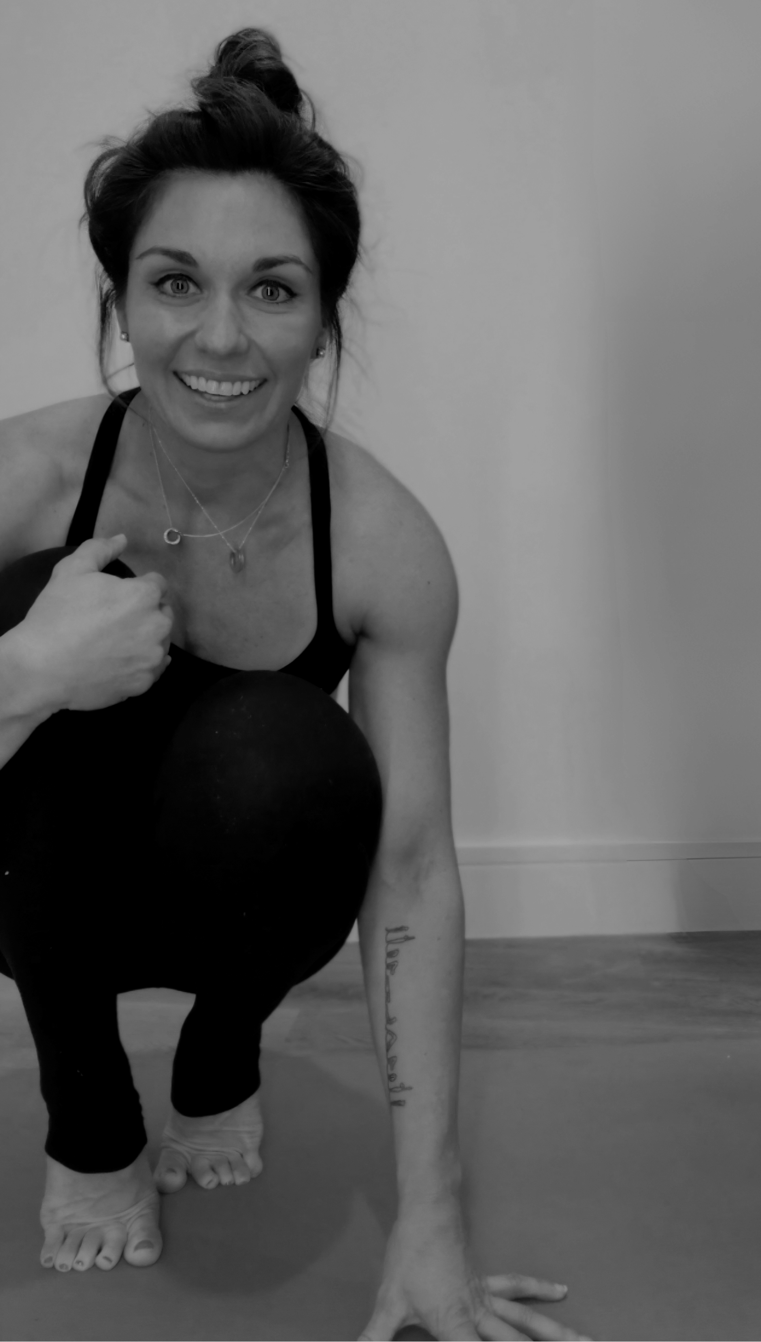 A woman with dark hair in a bun, wearing a sports bra and leggings, poses in a yoga pose on a mat indoors, smiling at the camera with a plain wall background.