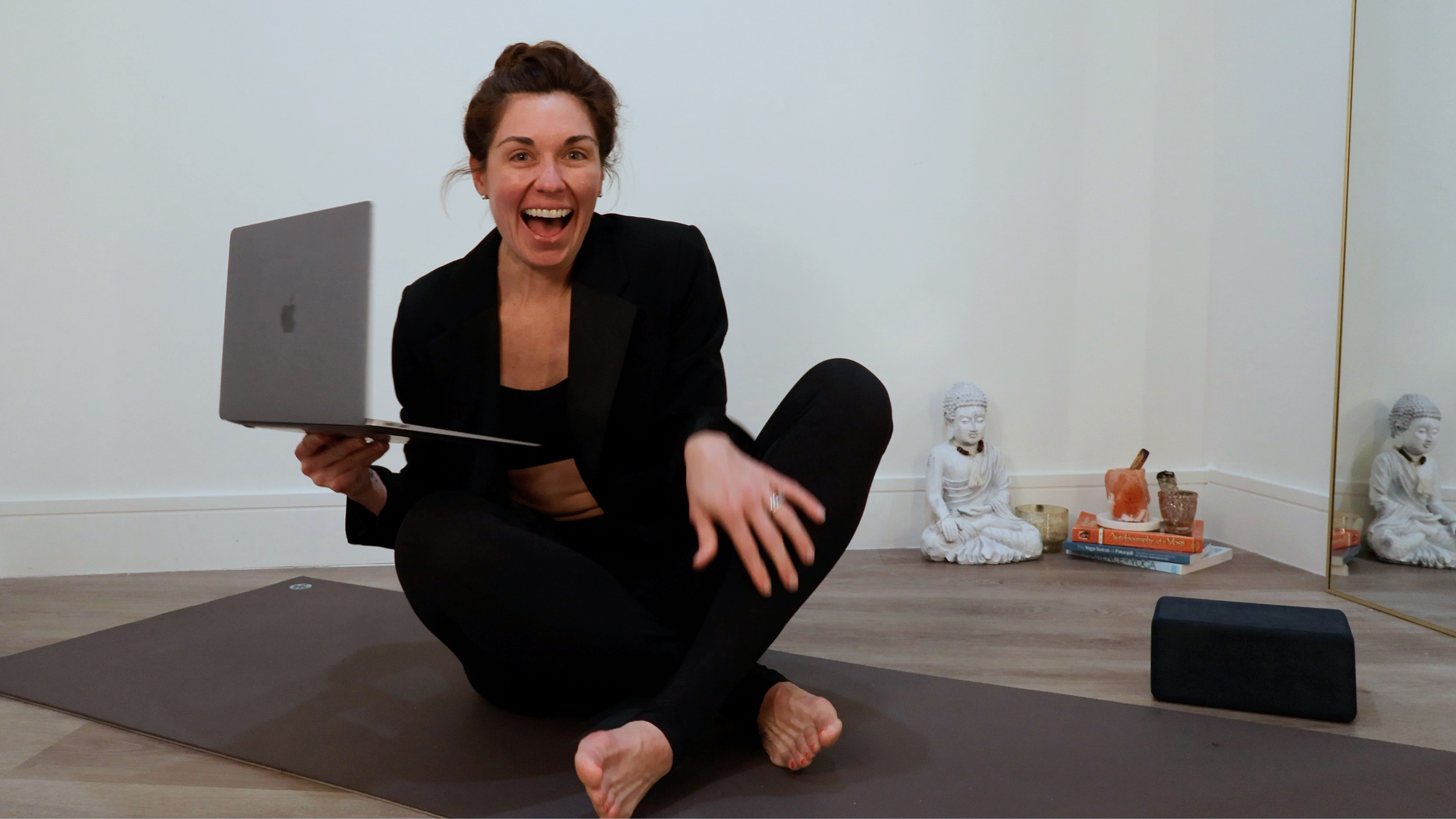 Brittney Coates Yoga Yoga, with dark hair in a bun, wearing a black blazer and black leggings, sitting on a yoga mat, holding a silver laptop, and smiling at the camera in a meditation or yoga space with Buddha statues and books in the background.