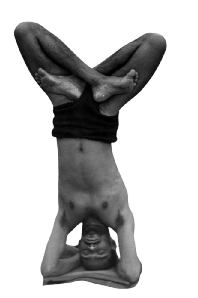 Upside-down shirtless man smiling, sitting cross-legged with hands behind his head, black background.