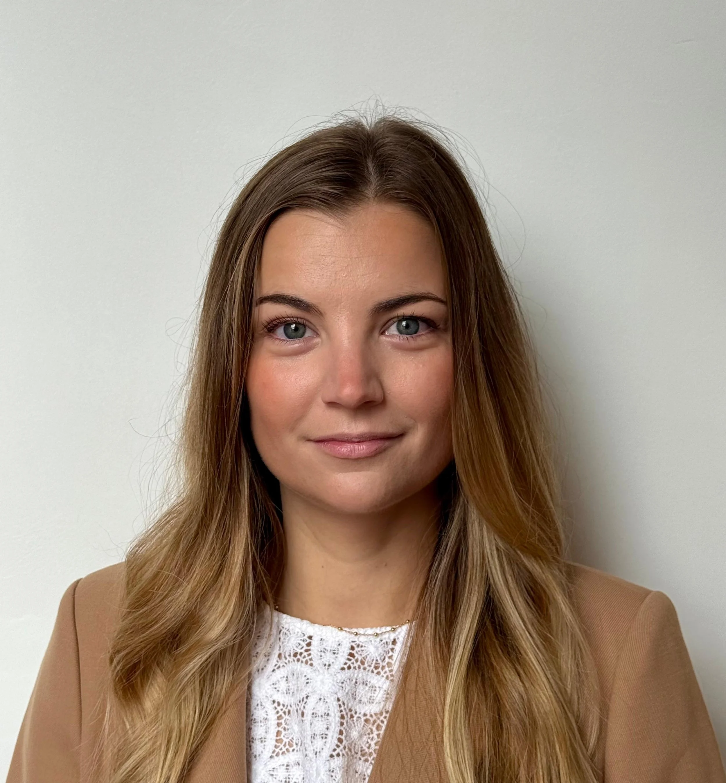Une femme avec des cheveux longs et bruns, portant un blazer beige et une blouse blanche en dentelle, souriant légèrement, photo prise devant un mur blanc.