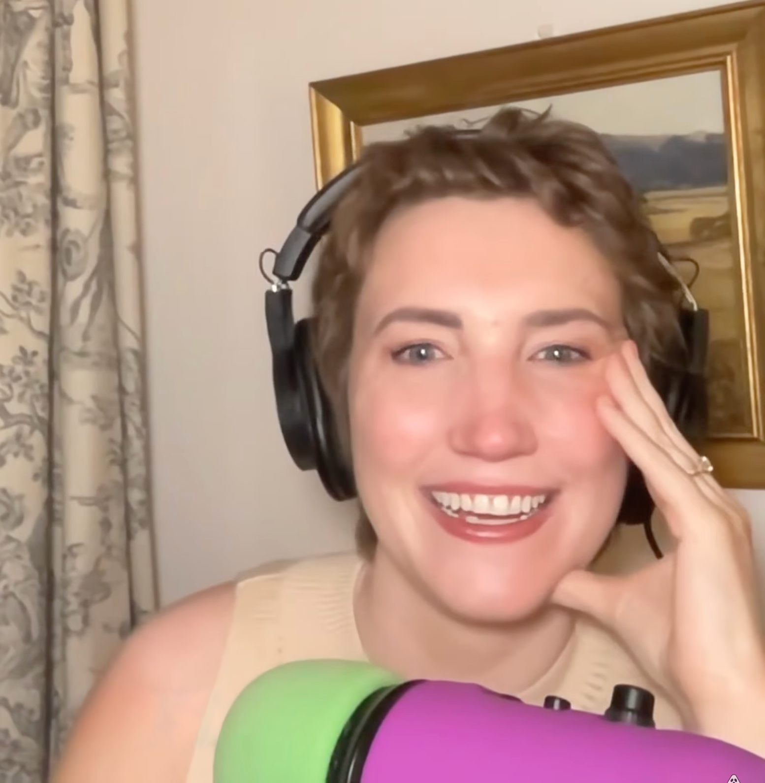 A woman with short curly hair wearing headphones, smiling and touching her face, sitting with a colorful microphone in front of her, in a room with patterned curtains and a painting on the wall.