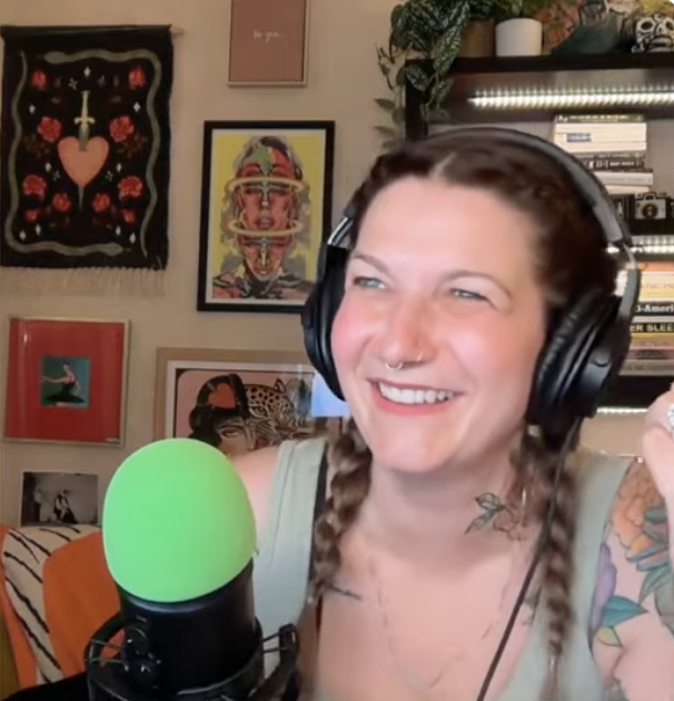 A woman with braided hair, wearing headphones, smiling in front of a microphone with a green windscreen, seated in a room with colorful artwork and books on a shelf in the background.