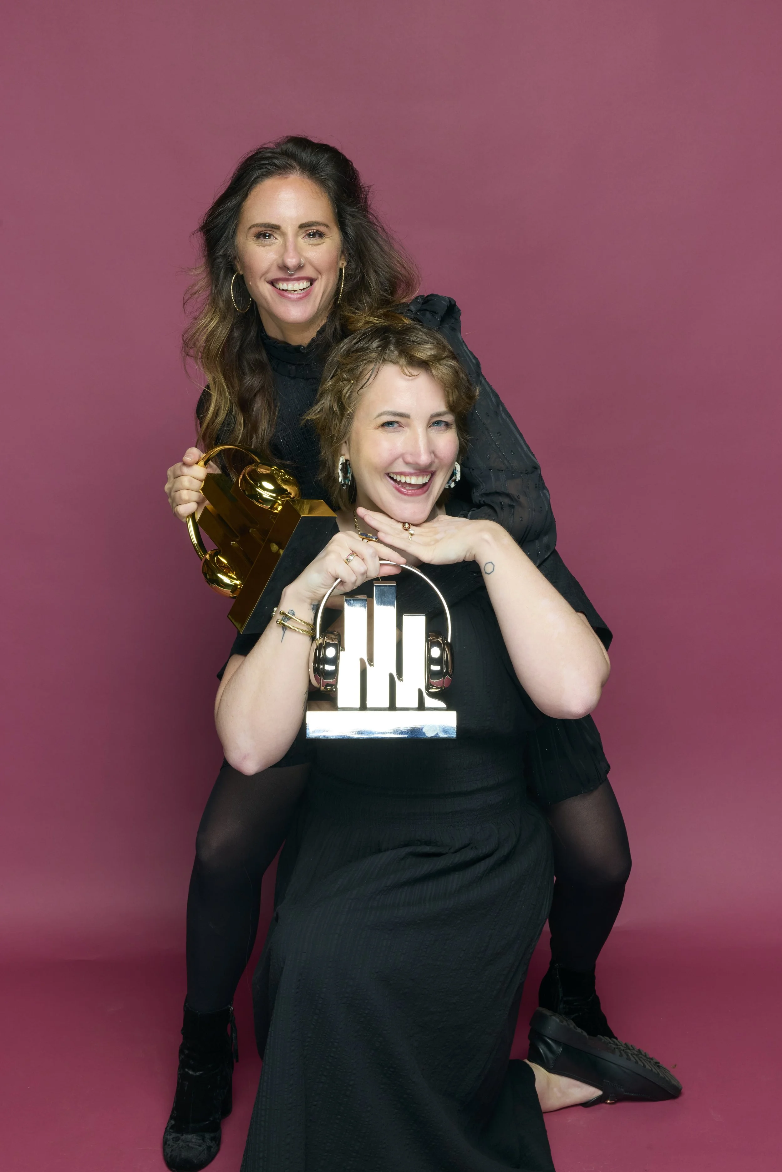 Two women, smiling and holding awards, standing against a pink background. One woman is sitting with headphones and a trophy, the other is standing behind her with a golden gramophone trophy.