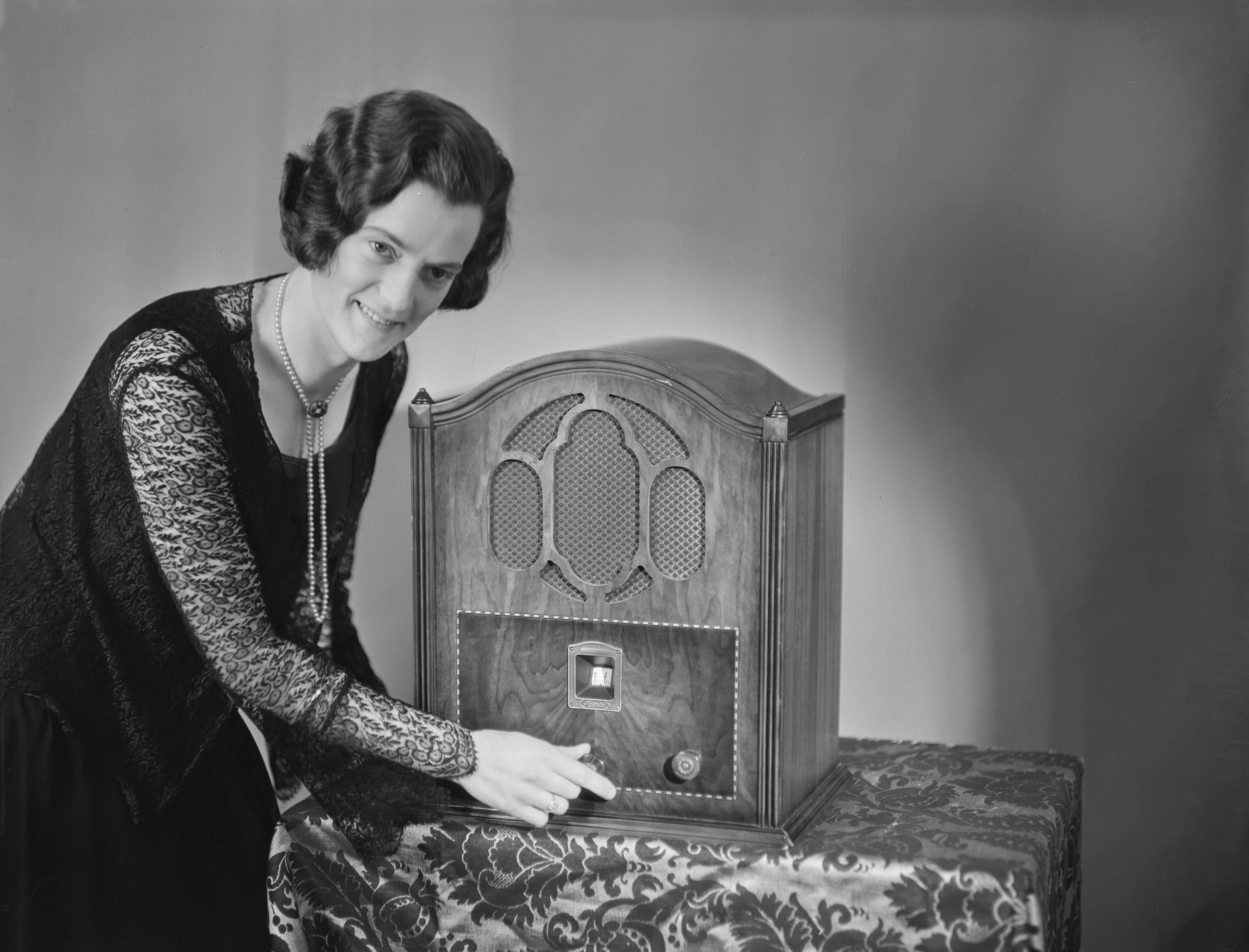 A woman dressed in vintage clothing, wearing pearls, poses with a wooden antique radio on a decorative table.