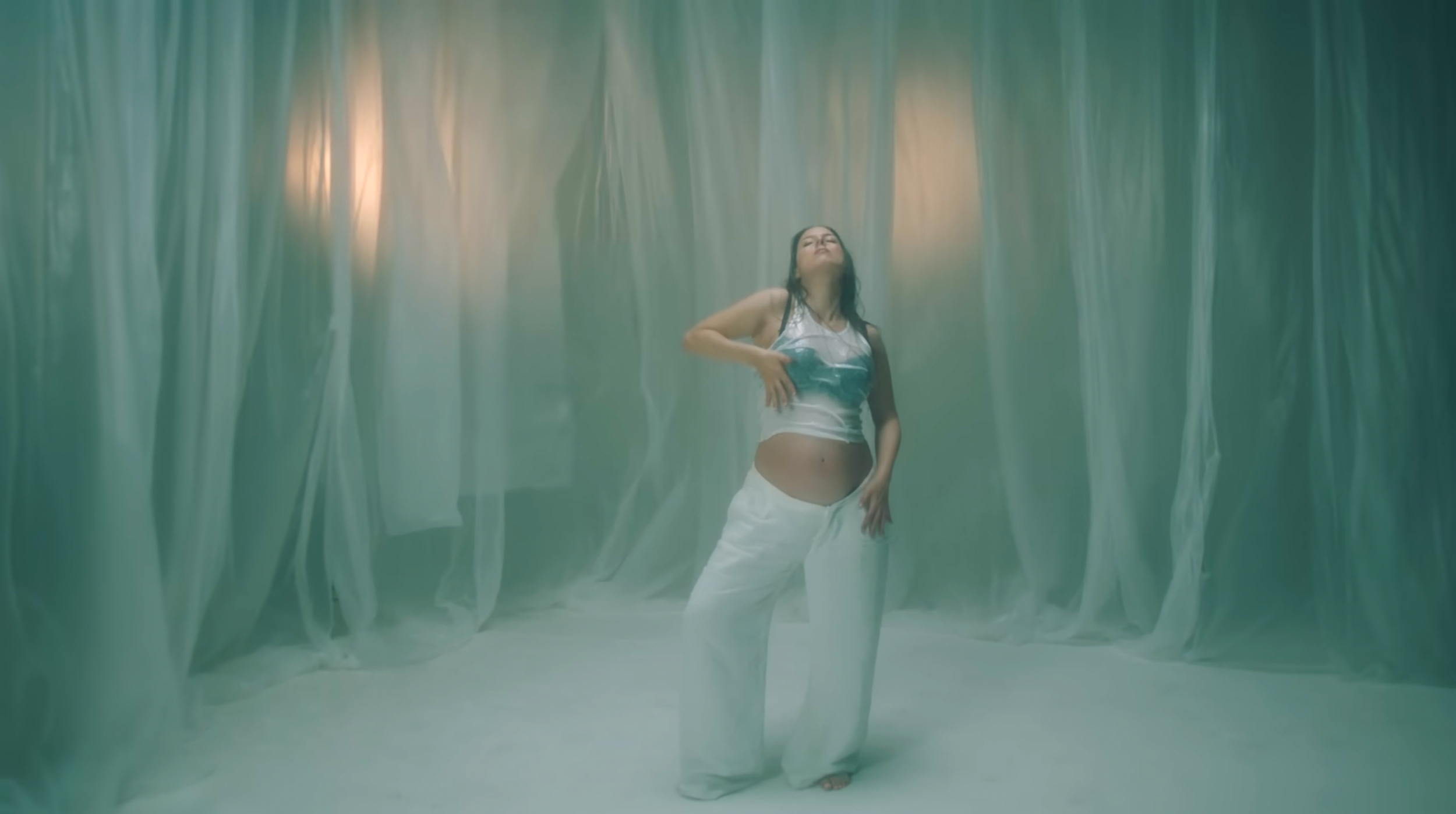 A woman standing in front of green curtains, wearing a shiny silver top and white wide-leg pants, with her eyes closed and one hand on her stomach.