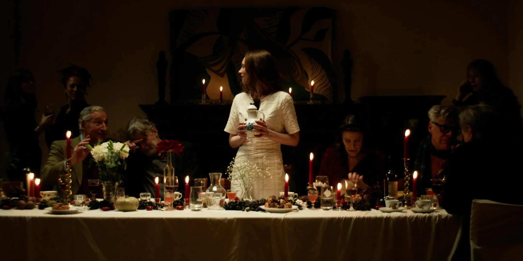 A woman in a white dress standing at a dinner table during a recorded or live celebration, possibly a Christmas or holiday gathering, with candles, flowers, and various foods, as other people sit and stand around her in a dimly lit room.