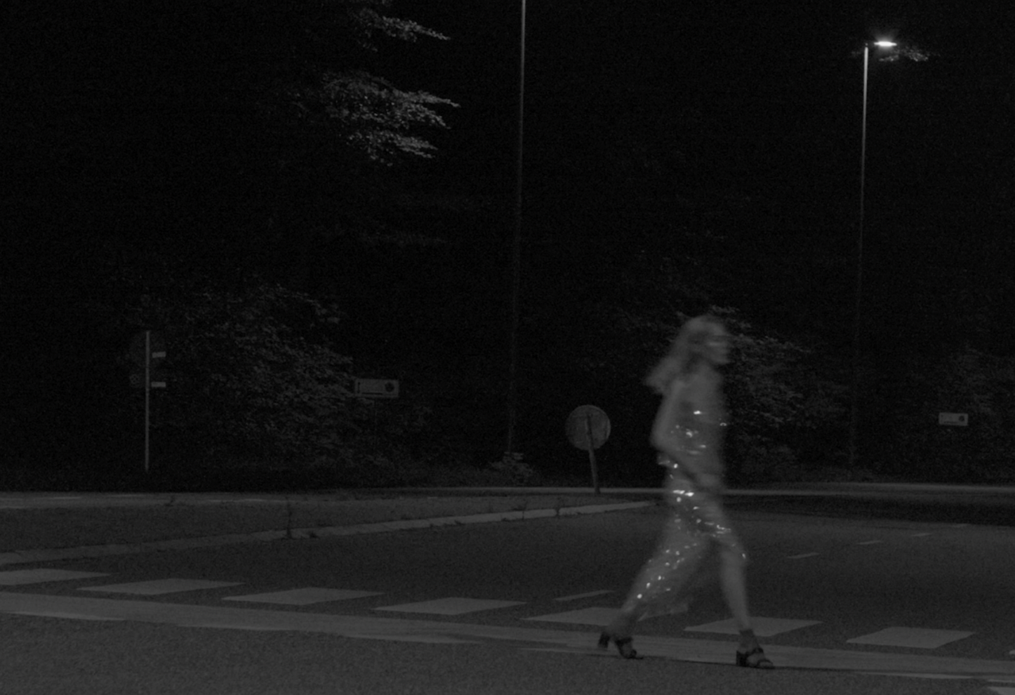 A woman walking across a crosswalk at night, dressed in a sparkly outfit, with streetlights illuminating the scene.