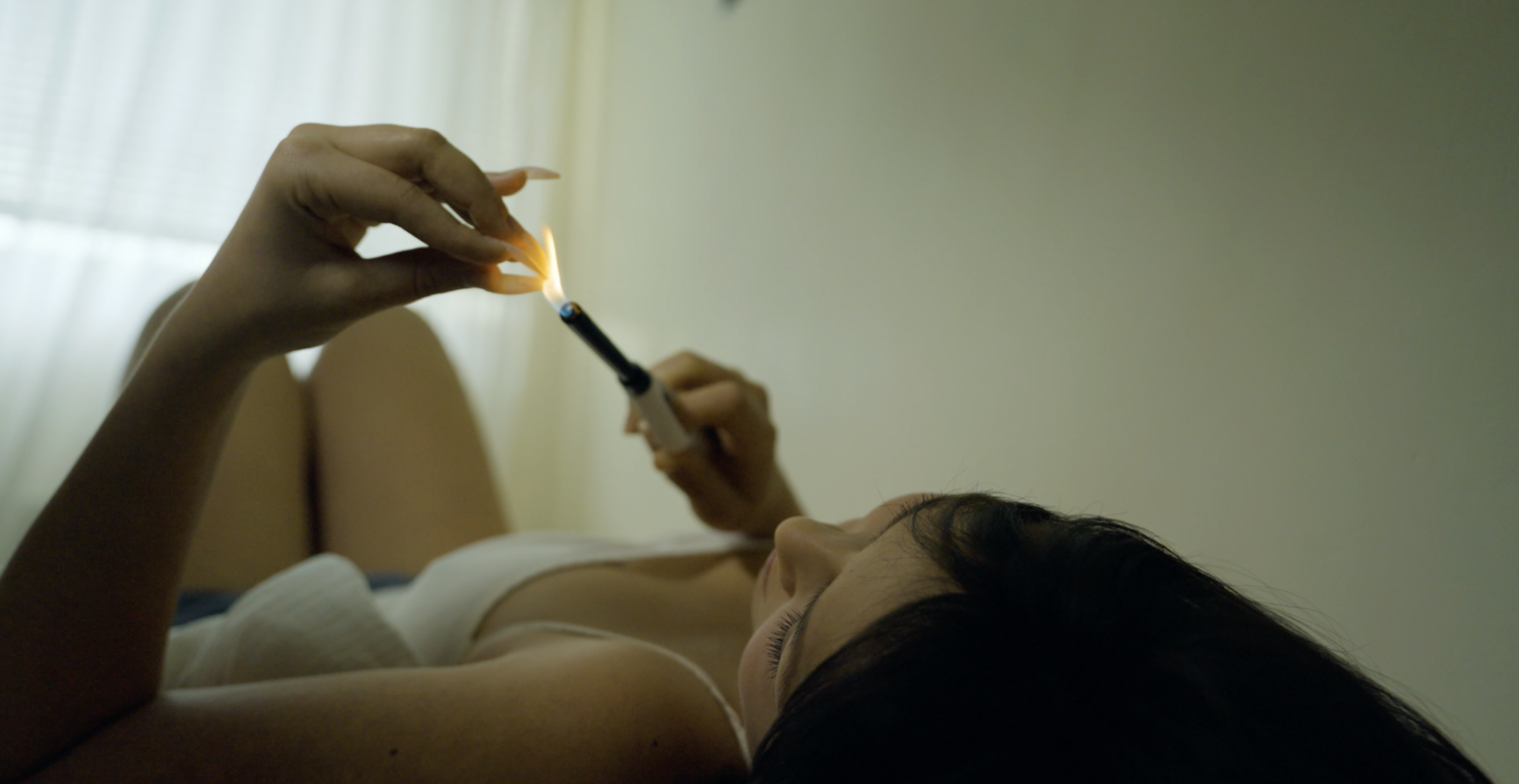 A woman lying in bed, holding a lit cigarette in one hand and a lighter in the other, with curtains in the background.