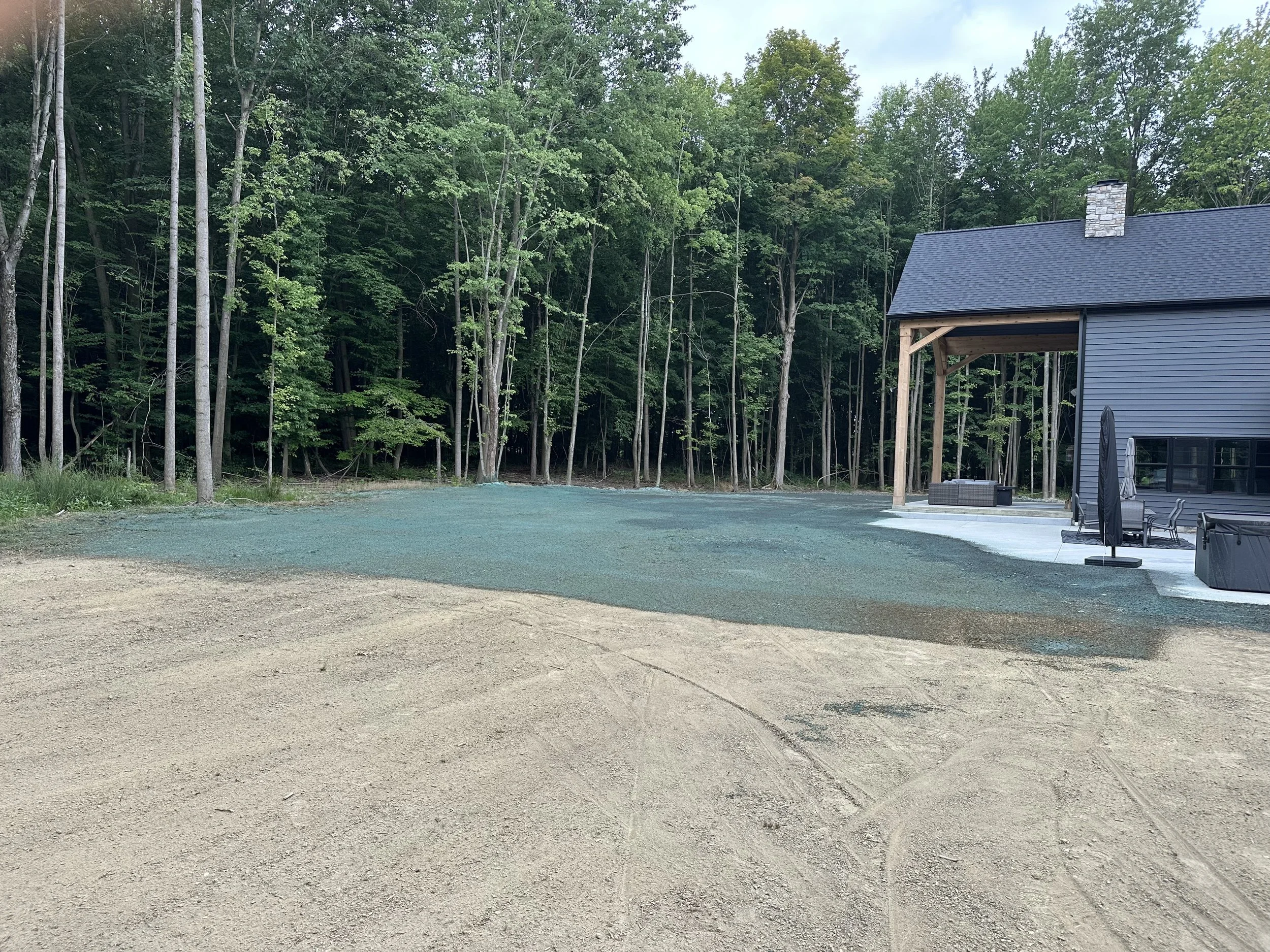 Partial view of a modern house with a covered patio area and outdoor furniture, set against a backdrop of a dense forest.